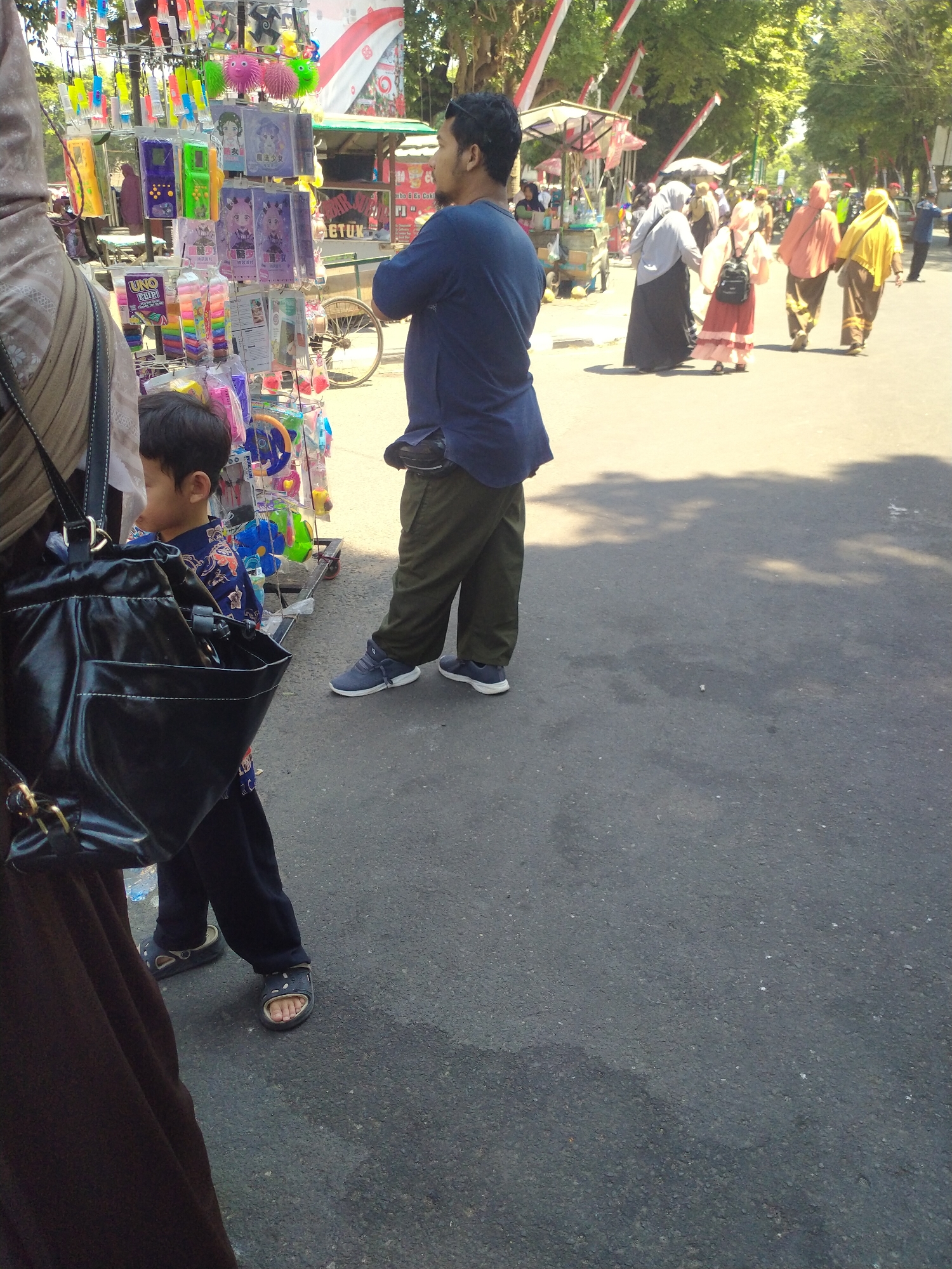 This image captures a street scene in Sukoharjo, Indonesia, likely during daylight hours due to the bright lighting and clear shadows. In the foreground, a person wearing a brown hijab and a dark brown garment is partially visible on the left. Beside them, a young boy in a blue and orange patterned shirt stands looking towards a display of colorful merchandise. The merchandise, displayed on a metal rack, includes various toys and novelty items such as small plastic figures, keychains, and what appear to be small electronic games.
To the right of the boy and the merchandise stand, a man wearing a navy blue shirt and olive green pants stands facing away from the camera, with his hands behind his back. He is wearing blue sneakers. The background reveals more of the street, with other people walking in the distance, some wearing hijabs and traditional Indonesian attire in various colors like peach, pink, and yellow. There are also stalls or shops visible further down the street, some with awnings. Trees line the right side of the street, providing shade and creating dappled light on the asphalt pavement. The overall atmosphere suggests a bustling marketplace or a street fair.