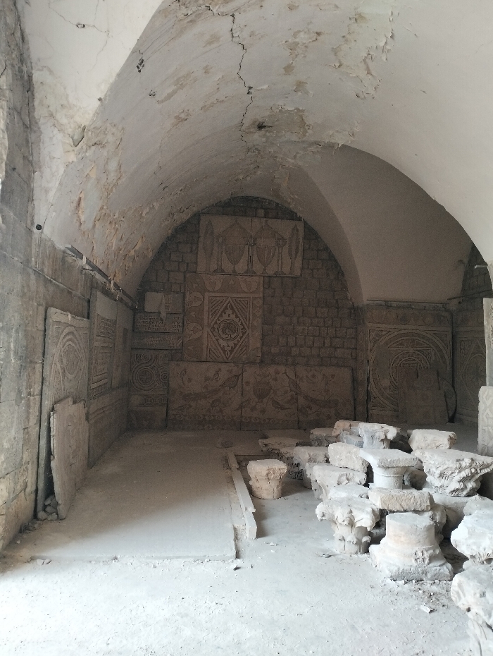 A weathered stone chamber features an arched ceiling with cracked paint and exposed brick. Carved stone panels adorn the walls, displaying intricate geometric and figurative designs. Scattered fragments of stone columns lie on the floor. The room's interior is bathed in diffuse light, highlighting the aging textures of the stone and the overall decay.