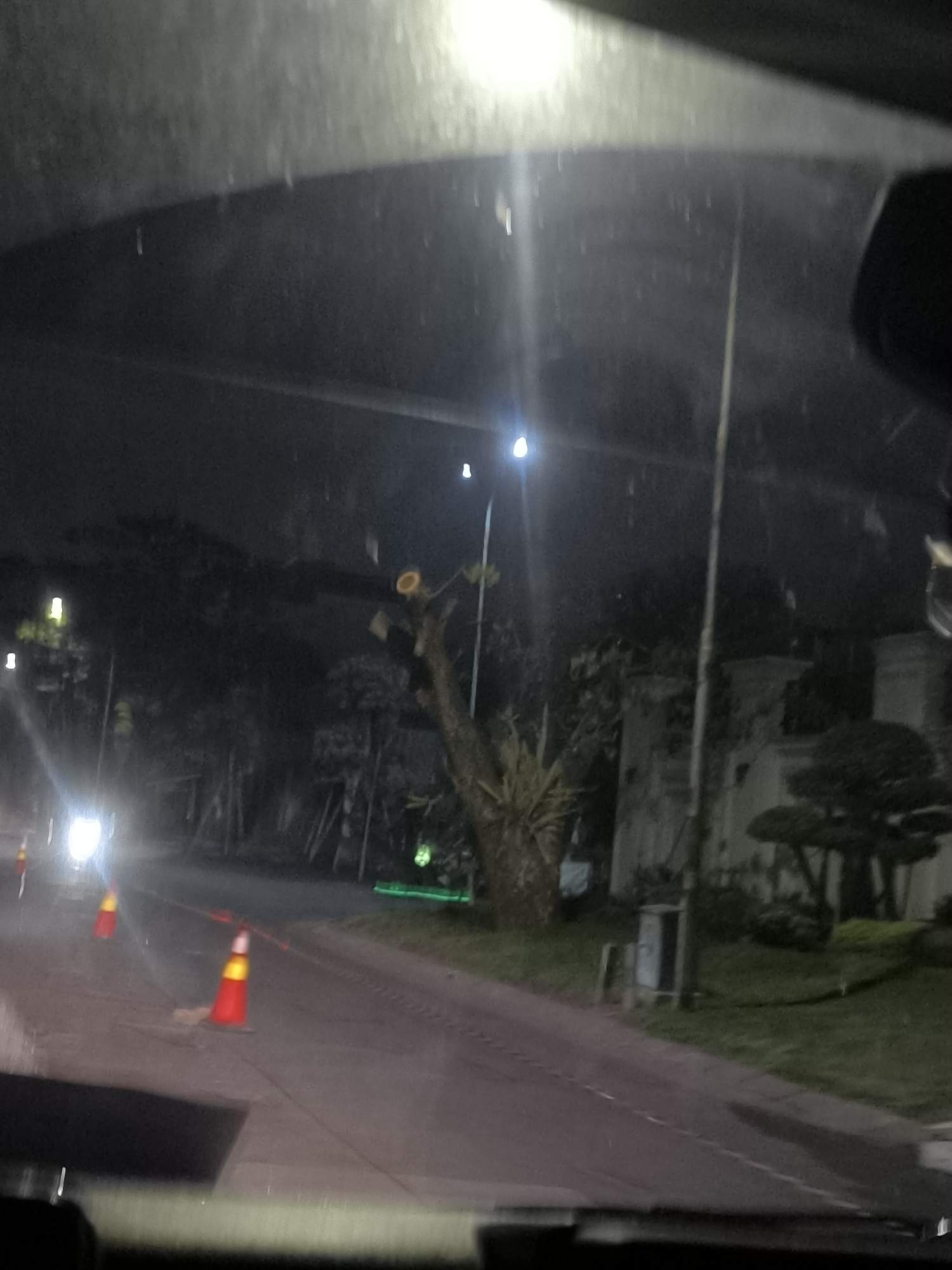 The image shows a nighttime view from inside a vehicle in Tangerang, Indonesia. A street is visible with traffic cones placed on it, suggesting roadwork or a temporary obstruction. Streetlights illuminate the scene, casting a glow on the wet pavement. Trees and buildings are dimly visible in the background. The overall impression is that of a quiet, urban street at night.