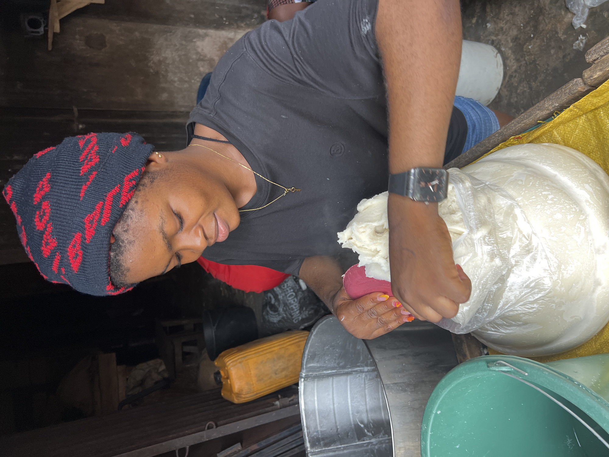 In Nkpor, Nigeria, a young woman wearing a black beanie with red patterns and a black t-shirt is actively engaged in preparing a large quantity of white, creamy foodstuff which is contained in a transparent plastic bag. Her hands, adorned with colorful nail polish, are shown in the process of scooping the foodstuff with a pink utensil and placing it into the bag. She is also wearing a watch on her left wrist.
The environment suggests an outdoor or semi-outdoor workspace, possibly a market stall or a kitchen area. Behind her, various items are visible: a large yellow jerrycan, a black bucket, a metal basin, and a green bucket. The background shows a dimly lit, rustic structure, possibly made of concrete or corrugated metal, with wooden elements. The ground appears to be dirt or concrete. A yellow tarp is laid out beneath the foodstuff bag. The overall impression is one of a local food preparation setting, where a woman is diligently working to process ingredients.