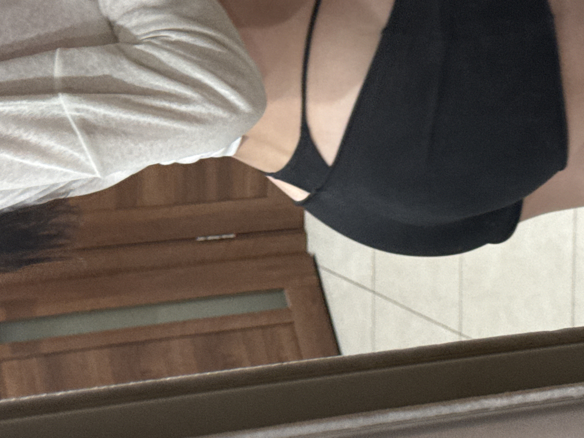 An individual is captured in a close-up, waist-down shot, indoors, likely within a residential setting. The subject is wearing a white, long-sleeved top and black shorts with a distinctive cut-out detail at the hip. The background features a wooden door with a frosted glass panel and a tiled floor, suggesting an interior space that could be a hallway or a room within a dwelling. The lighting appears to be ambient indoor lighting. There is no indication of this scene being located in Khimki, Russia.