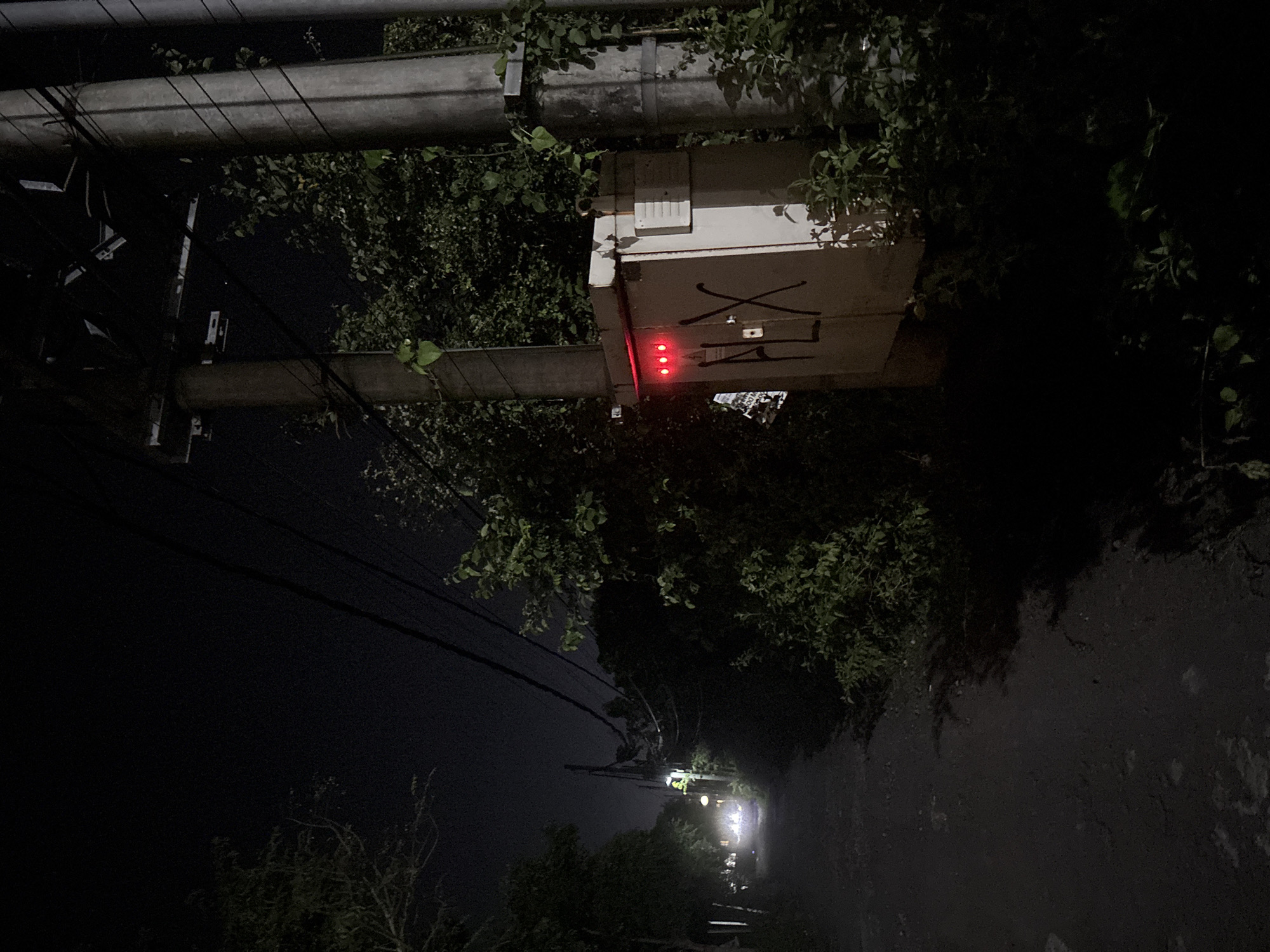 The image, taken at night in Ungasan, Indonesia, shows a utility pole with overgrown vegetation. A white electrical box is attached to the pole. The box has a black "X" painted on it, as well as other markings. Three red lights are illuminated below the markings. Power lines extend from the pole. A street with a light source in the distance is visible. The surrounding environment includes dark trees and a paved surface. There are no visible people.