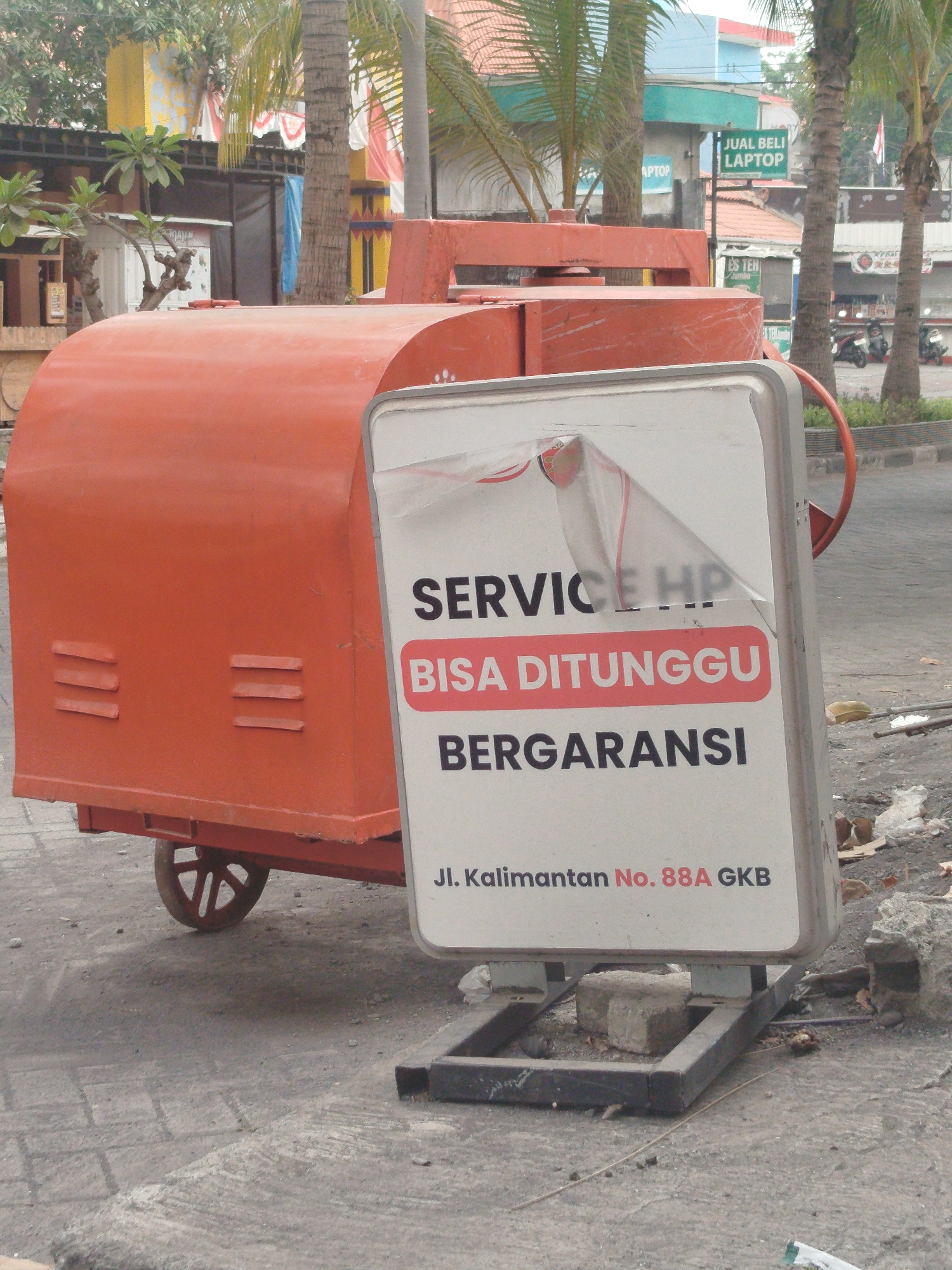In Yosowilangun, Indonesia, an orange industrial mixer is parked on a paved surface. To the right of the mixer is a white A-frame sign displaying text in Indonesian. The sign reads, "SERVICE HP BISA DITUNGGU BERGARANSI" and below that, "Jl. Kalimantan No. 88A GKB". A torn piece of clear plastic sheeting is partially covering the top of the sign, obscuring some of the text. The background shows palm trees, buildings, and parked motorcycles.