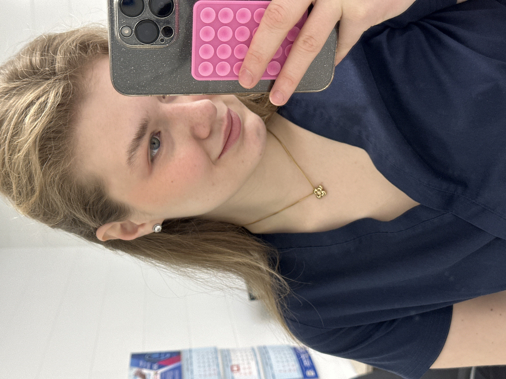 A person is taking a selfie indoors. The person has light blonde hair and fair skin, wearing a navy blue shirt. A gold necklace with a four-leaf clover pendant is visible. The phone in use has a gray case with a pink pop-it attached to the back. An earring is seen on the person's left ear. A calendar is visible in the background, indicating that the location may be in Kazan, Russia.