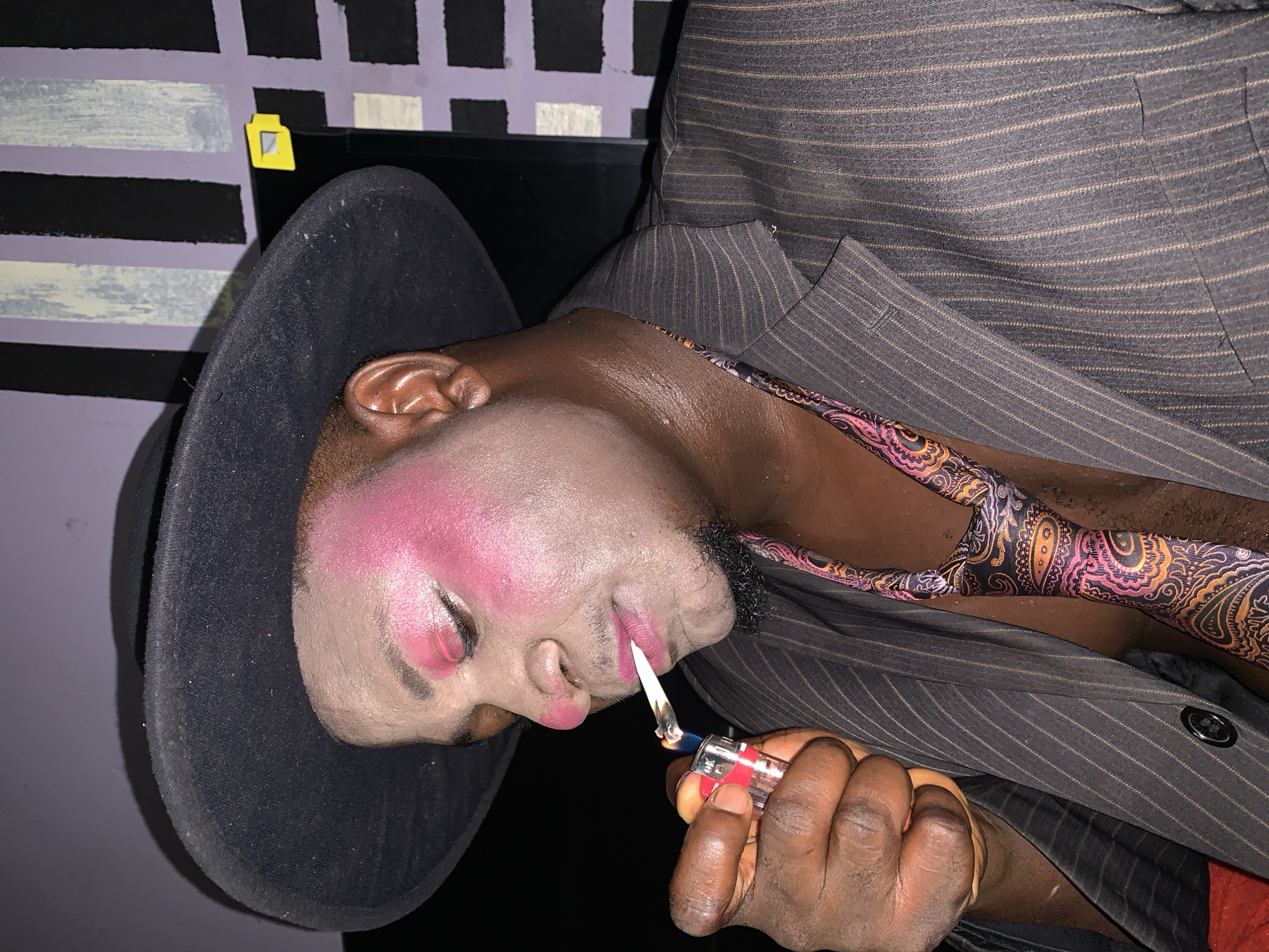 A person is shown in a close-up shot, wearing a black fedora hat, and a pinstripe suit jacket over a colorful paisley tie. Their face is adorned with prominent pink blush on the cheeks and eyes, and pink lipstick. They are in the process of lighting a cigarette with a clear lighter, which is held in their right hand. The background appears to be a wall with a striped pattern of black and light purple. The scene is illuminated by artificial light, creating shadows and highlighting the makeup and textures of the clothing. The location is identified as Akwanga, Nigeria.
