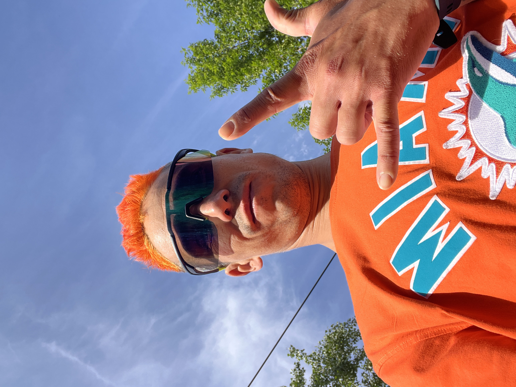 A man is pictured from a low-angle perspective against a blue sky with scattered clouds and green tree foliage in the background. He is wearing orange athletic sunglasses and an orange t-shirt that features the word "MIAMI" in blue lettering and a logo depicting a marine creature, possibly a dolphin. The man’s hair is dyed bright orange. He is making a gesture with his right hand, extending his index finger. The location is the city of Chaikovsky, Russia.