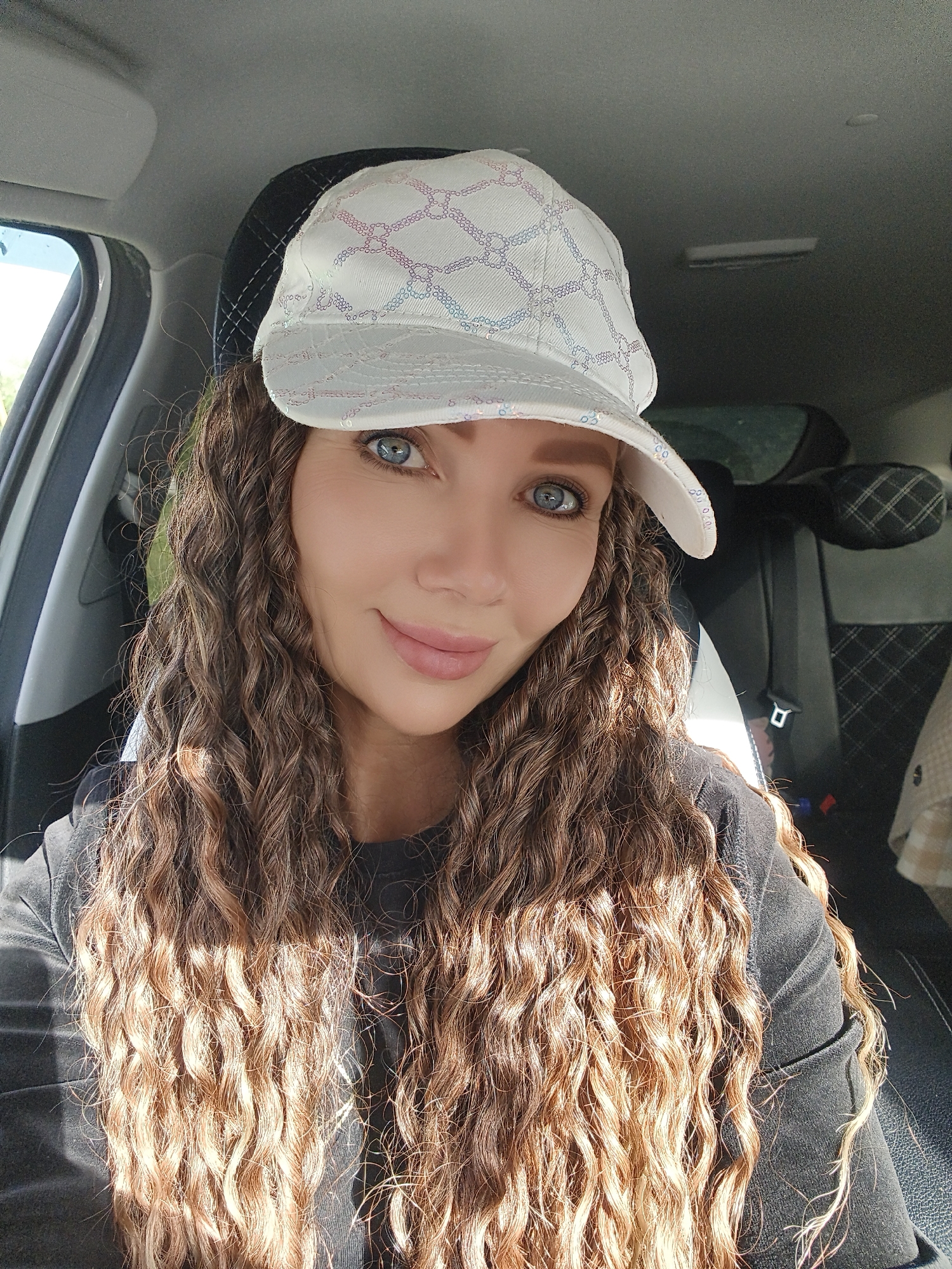 A woman with curly brown hair and blue eyes, wearing a white baseball cap with a patterned design and a black t-shirt, is seated in the driver's seat of a car. The interior of the car is visible, including the headrest and seatbelt. The lighting suggests it is daytime, possibly sunny. The woman is looking at the camera and smiling. The background outside the car window is blurred, showing some greenery.