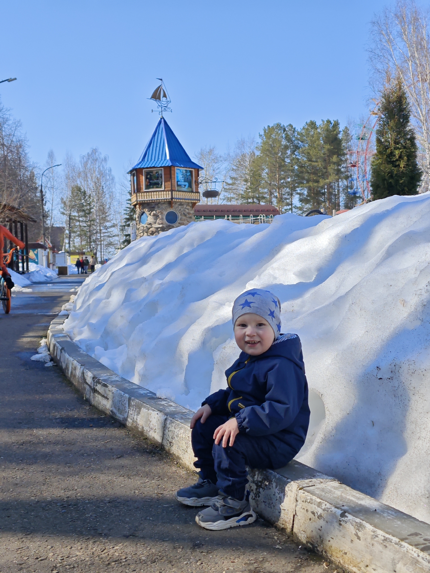 The image shows a young child sitting on a low concrete wall in the city of Chaikovsky, Russia. A large pile of snow is behind the child. In the background is a decorative tower with a blue roof and a sailboat weather vane, along with evergreen trees and a glimpse of a Ferris wheel. A person on a bicycle and other figures are visible in the distance. The sky is clear and blue, suggesting a sunny day.
