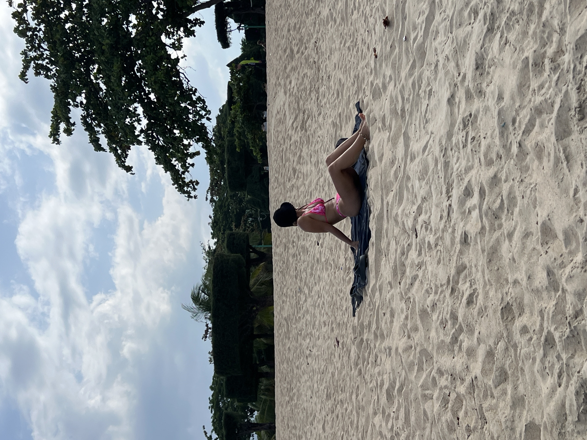 The image depicts a daytime beach scene in Nha Trang Ward, Vietnam. The foreground and midground consist of light beige sand, showing ripples and shallow indentations. In the midground, a female individual is seated on a dark grey towel laid directly on the sand. She is positioned with her back partially to the viewer, facing towards the right side of the frame, and resting on her forearms. She wears a pink two-piece bikini and a dark-colored bucket hat. An anklet is visible on her right ankle. A pair of dark-colored flip-flops or sandals are on the sand near the towel.
In the background, a line of green foliage and trees is visible. Several distinct trees exhibit uniform, rectangularly pruned canopies atop clear trunks, resembling hedges on stilts. Further back and to the right, a large tree with dense, irregularly shaped green foliage partially extends into the upper right corner of the frame. To the far right, near the tree line, a small, indistinct human figure wearing a bright yellow-green shirt is visible standing. The sky above is partially overcast with scattered white and light grey clouds against a light blue backdrop.