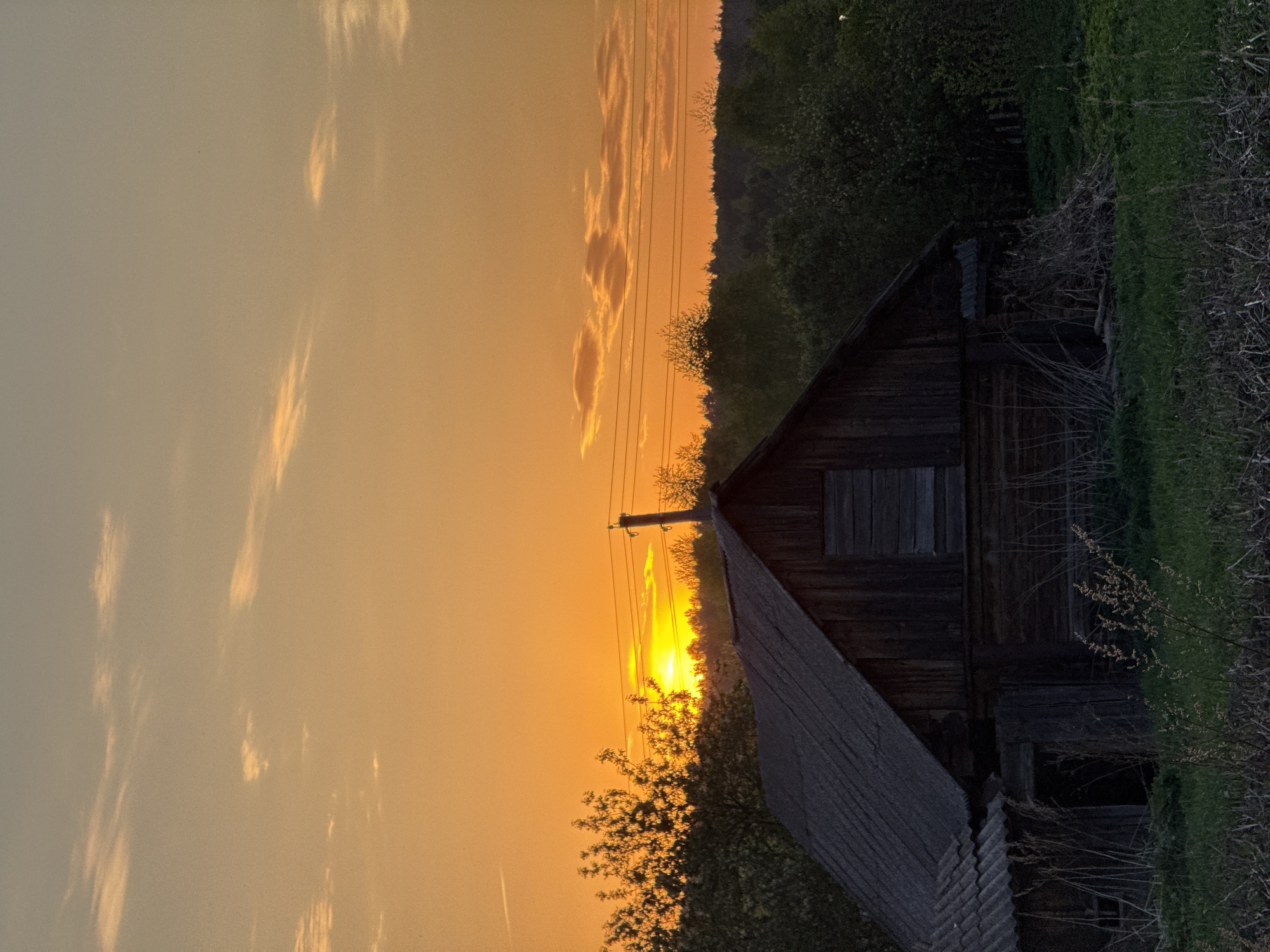 The image depicts a sunset scene in Nizhnyaya Chermoda, Russia, with a dilapidated wooden house in the foreground. The sun is setting, casting an orange glow across the sky and highlighting wispy clouds. Power lines and trees are silhouetted against the bright sky. The house shows signs of age and disrepair, with boarded-up windows and a weathered roof. Overgrown vegetation surrounds the building.