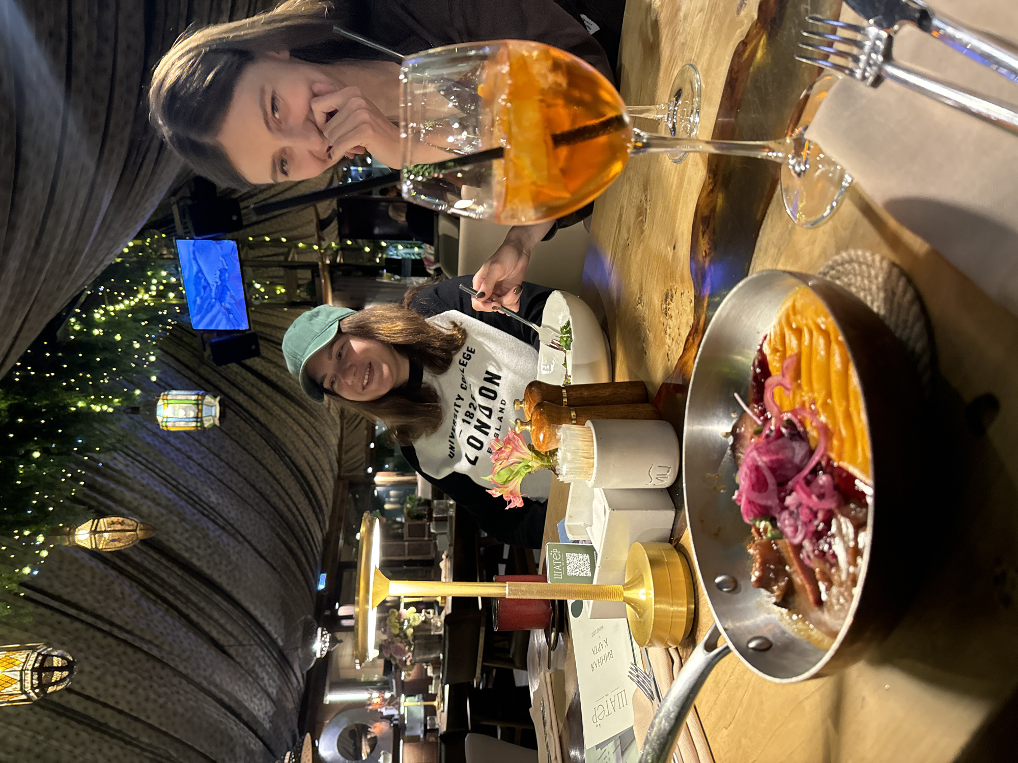 Two women are seated at a wooden table in a dimly lit restaurant in Moscow, Russia. One woman in a green baseball cap and a white sweater that reads "University College London" is smiling at the camera while holding a fork in her left hand, ostensibly eating a salad. The second woman is looking at the food on the table, with her hand partially covering her mouth. A cocktail with a straw and orange slice sits to her right. The table is set with silverware, a napkin, a wine list, condiments, and a dish featuring a colorful mix of pumpkin and red onions. The restaurant has a unique, textured roof, with lights and hanging lamps adding to the ambiance. A blue screen can be seen in the background.