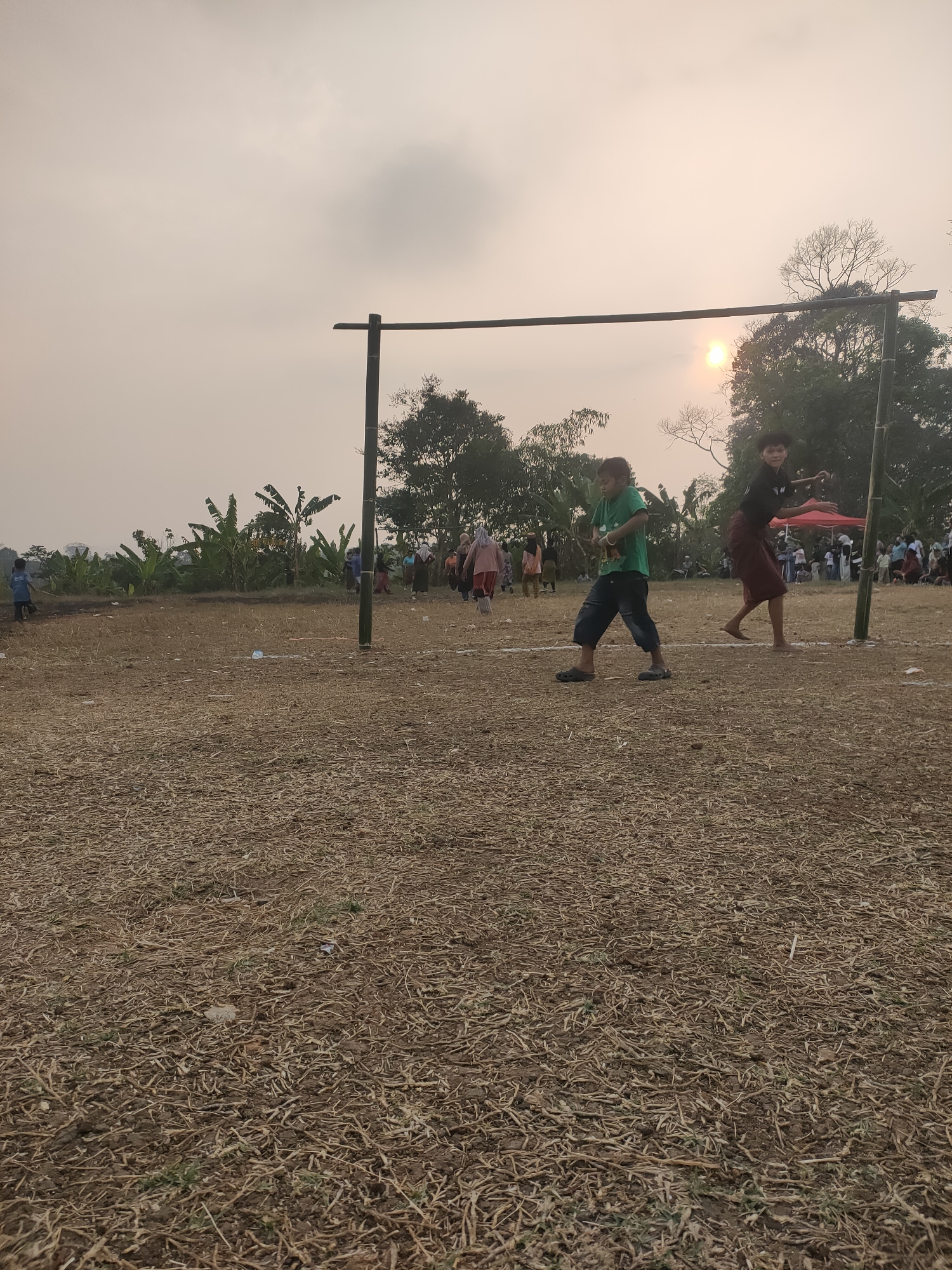 This image depicts an outdoor scene, likely a field or open ground, with a rustic wooden goalpost structure visible in the center. The ground is covered in dry, straw-like debris and dirt. Two young individuals are the prominent subjects in the foreground. One, wearing a green t-shirt and dark pants, is in motion, possibly playing a game. The other, in a dark shirt and maroon pants, is also in motion, appearing to be mid-action.
In the background, a crowd of people is gathered, some standing and some in motion. A red tent is visible in the distance. Trees and foliage, including what appear to be banana trees, are present on the left side of the frame. The sky is overcast with a hazy sun visible through the clouds, suggesting either dawn or dusk. The overall impression is of a community event or activity taking place in a rural or semi-rural setting. The presence of a goalpost implies a sporting event or game. The context of Subang, Indonesia, suggests this could be a local festival, a school sports day, or a casual gathering.