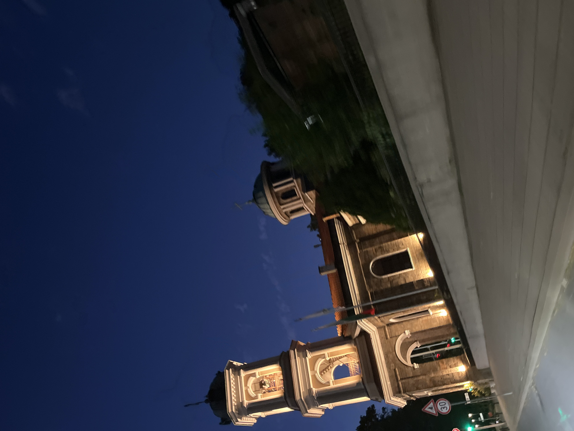 The image shows a building that appears to be a church or a cathedral, with ornate architecture and illuminated windows. It has two bell towers, one of which is more prominent in the foreground, featuring arched openings and bells. The building's facade is detailed with columns and decorative elements. A dome-like structure is visible on top of the main building.
The scene is set against a deep blue twilight sky, suggesting it is evening. The lighting on the building indicates it is illuminated, likely for nighttime viewing. Trees and greenery are visible in the background, and a road or elevated walkway runs alongside the building. Traffic lights are visible, one showing green. There are also some traffic signs visible, including a circular sign with "30" inside and a triangular warning sign.
The overall impression is one of a peaceful evening scene in an urban or semi-urban setting. The image is oriented sideways. There is no visible text on the building itself, but the traffic signs contain numerical and symbolic text. The location is identified as Plovdiv, Bulgaria.