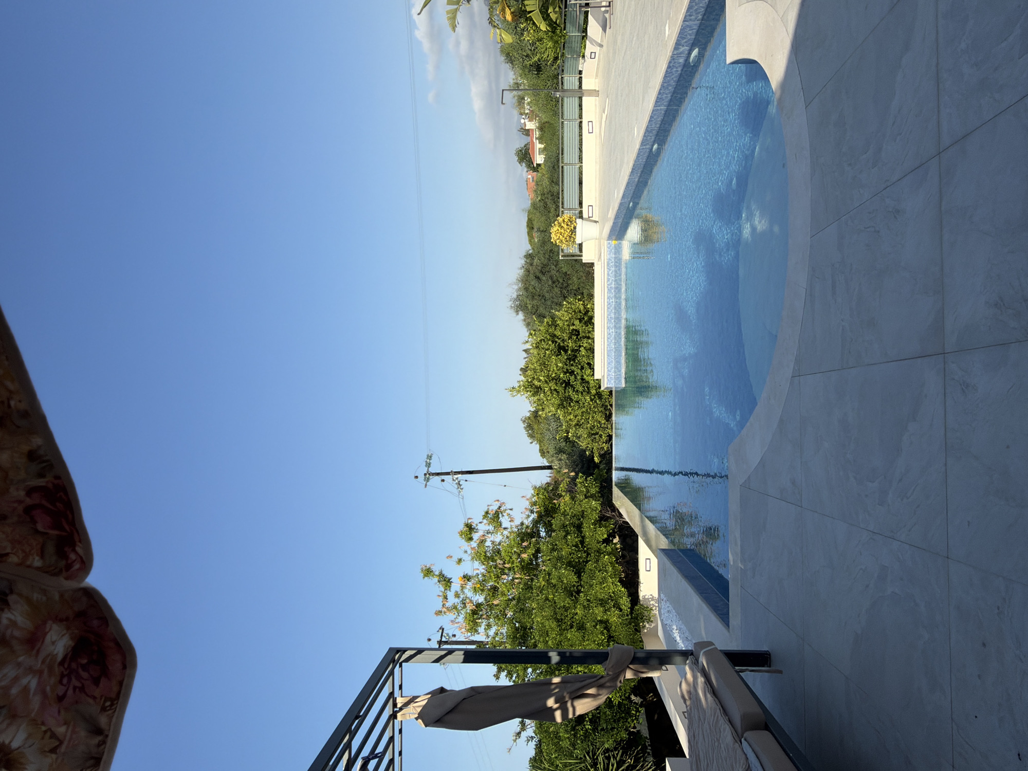 A bright, sunny day is captured in this image of a resort-style swimming pool area. The pool itself is rectangular with a curved section, filled with clear blue water that reflects the sky and surrounding trees. The pool is surrounded by a light-colored tiled deck. To the left, a portion of a lounge chair with a beige cushion is visible under a dark metal pergola. A folded beige umbrella hangs from the pergola. Lush green trees and some flowering bushes are in the background, along with power lines and poles. In the distance, houses with red roofs are nestled amongst the trees. The sky is a clear, bright blue, with a few wispy clouds towards the right side of the frame. The overall impression is one of a well-maintained, tranquil outdoor living space, likely in a warm climate such as Kyrenia, Northern Cyprus.