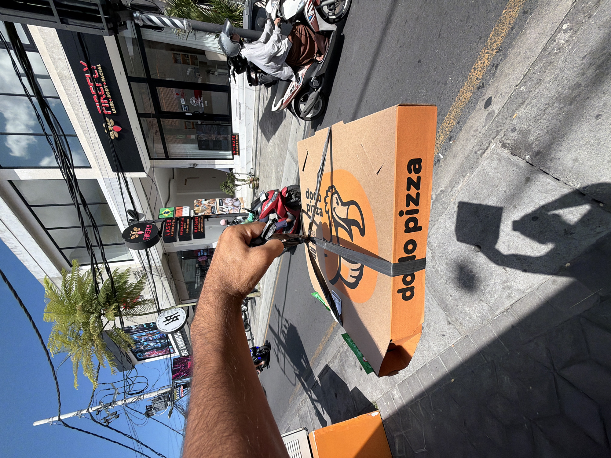 The image presents a bright, sunlit street scene in North Kuta, Indonesia, viewed from an elevated, slightly overhead perspective. In the foreground, a person's bare forearm and hand are visible, using scissors to cut a black securing strap on a large, orange-brown pizza box. The box is prominently branded with "dolo pizza," featuring a stylized chicken head logo.
The background reveals a bustling urban environment. On the asphalt road, a person wearing a helmet and light attire is riding a white and red scooter. The street is lined with commercial buildings featuring various storefronts. One notable business is "FIREFLY LASER + BODY + FACIAL," displayed on a black awning and a round sign. Another sign lists services including "LASER HAIRREMOVAL," "HYDRAFACIAL WITH DERMALOGICA," and "MASSAGE (WOODTHERAPY, LYMPHATIC, RELAX)." Other partial signs refer to "INTIMATE BROWSING," "RIE TANNING," and "TEMPORARY TATTOO."
Numerous power lines and utility poles crisscross the clear blue sky above the street. Strong shadows cast by buildings and objects, including a person's shadow, indicate the bright midday sun. A green, fern-like plant hangs near one of the buildings. The activity suggests the opening or preparation of a pizza delivery on a typical sunny day in the region.