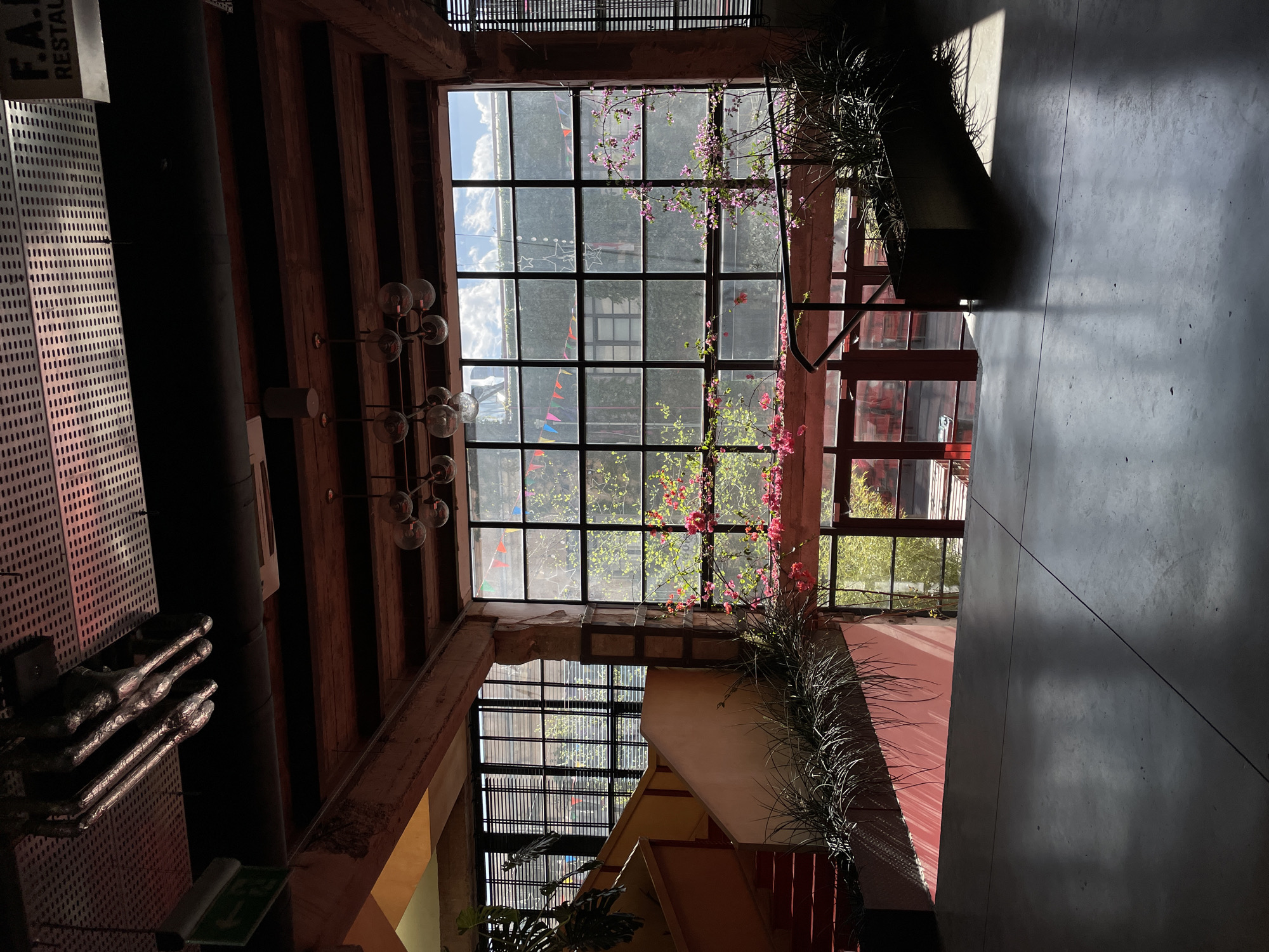 This media file captures an interior view of a modern, multi-level building, likely a restaurant or a public space, looking upwards. The scene is dominated by a large, grid-patterned window or skylight at the top, through which a bright, partly cloudy blue sky and green trees are visible. Pink flowering vines with numerous blossoms elegantly drape across the window frames and hang downwards, adding a vibrant natural element to the industrial aesthetic.
On the left side of the image, a perforated metal wall features what appear to be abstract silvery sculptures or light fixtures. Above this, a partially visible sign reads "F.A.I RESTAU", suggesting a restaurant. A large, dark pipe or ventilation duct runs horizontally across the upper left. A modern chandelier with multiple spherical glass lights hangs from the ceiling, which is composed of exposed wooden beams and brickwork.
The right side of the building showcases concrete walls and floors, illuminated by strong sunlight that creates dramatic, elongated shadows. Long, spiky green plants are arranged in planters along ledges. Below the main window, a complex architectural design includes a staircase or ramp with geometric shapes and another grid-patterned window looking out onto more greenery and colorful triangular flags or bunting. A green emergency exit sign with a running person pictogram is visible in the lower left.
The natural light suggests it is daytime, likely morning or afternoon, with clear weather outside. No people are visible, indicating a quiet moment. The overall atmosphere is modern and airy, with a blend of industrial materials, natural light, and lush greenery. The location context provided is Tbilisi, Georgia.
