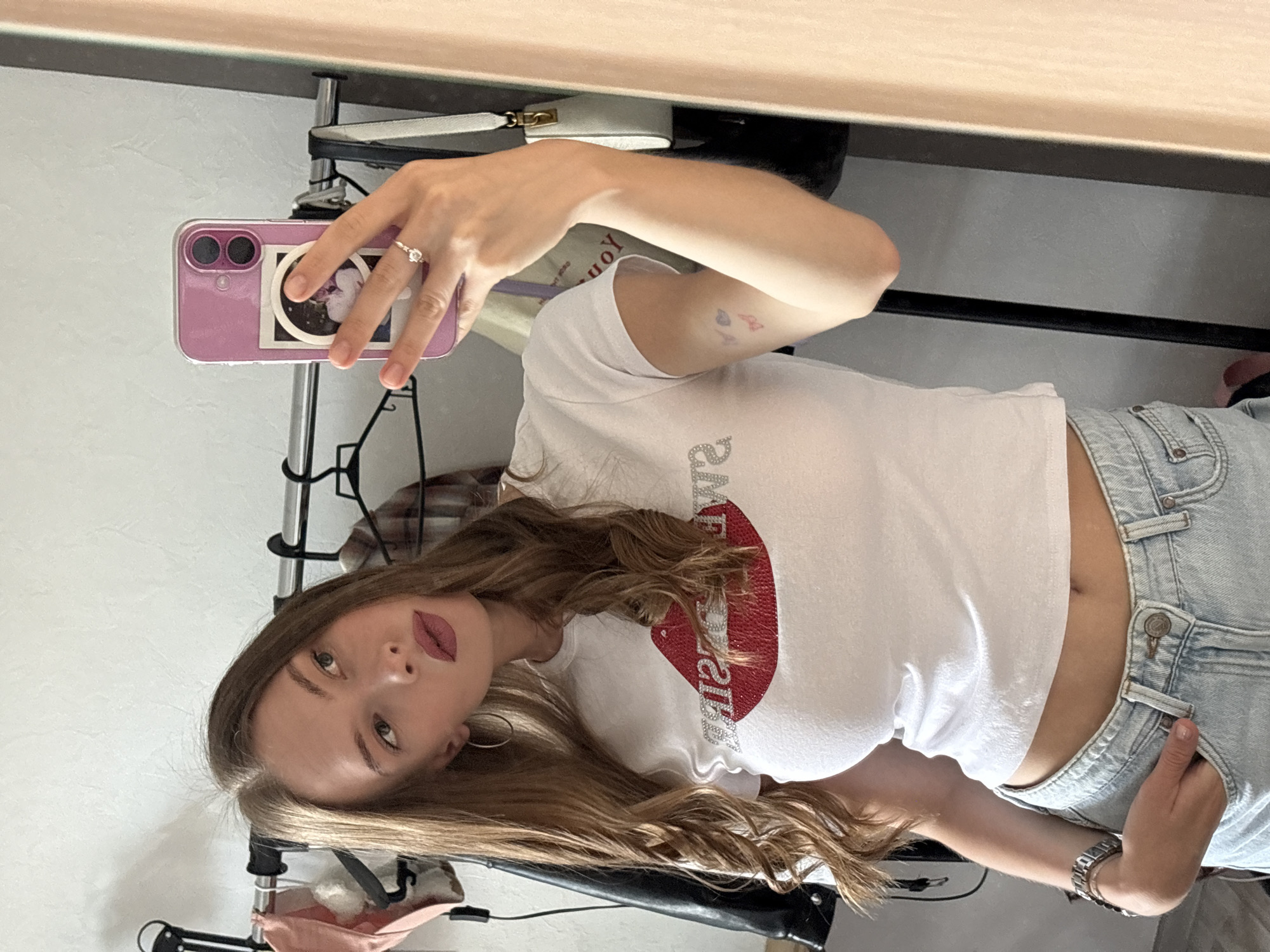 A young woman is taking a mirror selfie. She is wearing a white t-shirt with a large red sequined lip design and light-wash denim jeans. She has long, wavy brown hair, bold pink lipstick, and hoop earrings. A silver ring is visible on her left hand, and a watch is on her left wrist. She has small tattoos on her upper right arm, one resembling a butterfly and another a heart. She is holding a pink phone in her right hand, which has a sticker on the back. The background appears to be an indoor setting, possibly a room, with a textured white wall and a reflective surface, likely a mirror, where the selfie is being taken. The lighting suggests daytime.