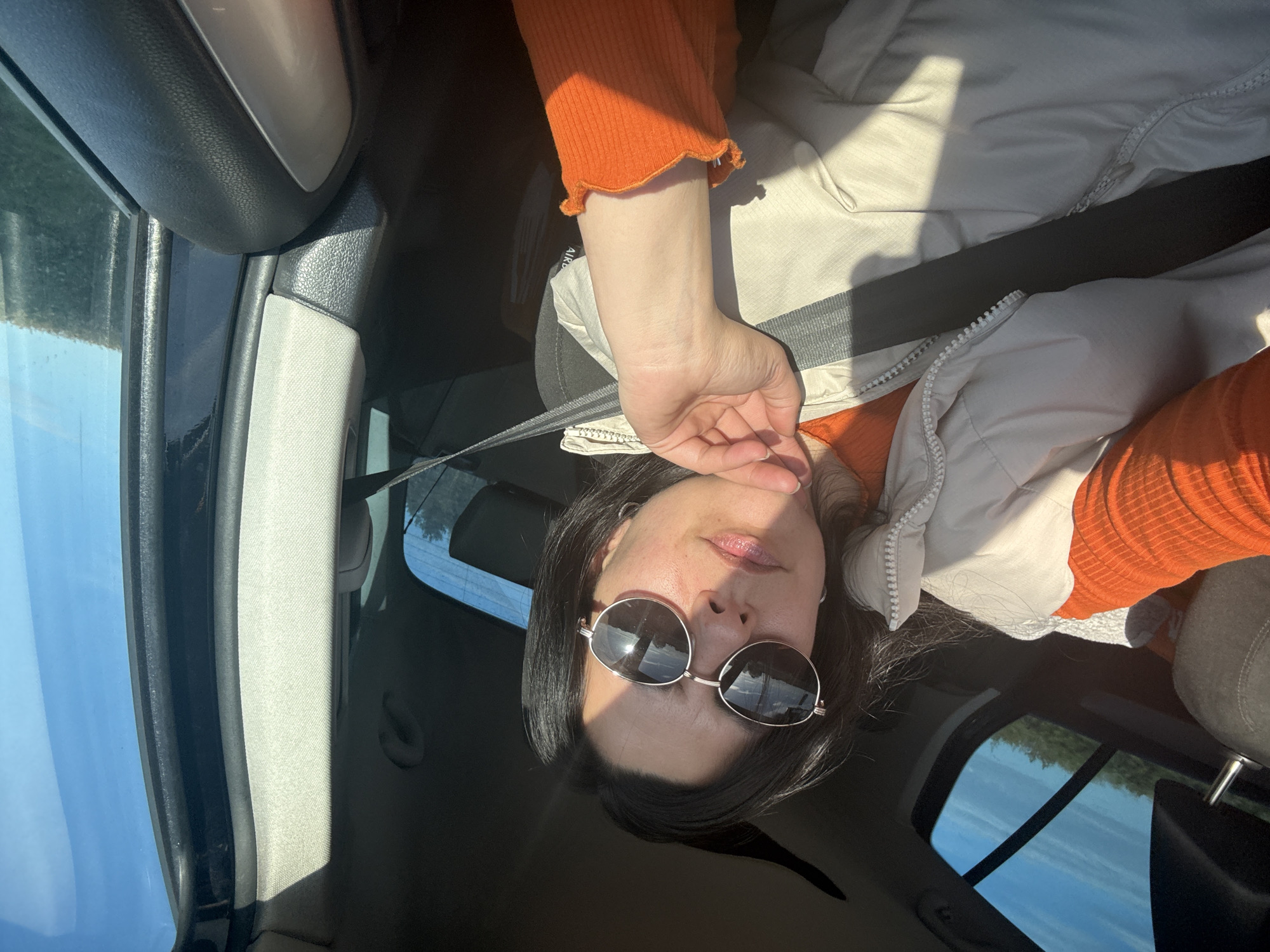 A person with dark hair, wearing sunglasses and a seatbelt, is in a car. They are wearing an orange top and a light-colored vest. The location is Breckland District, United Kingdom, suggested by the reflection of UK buildings on the sunglasses.