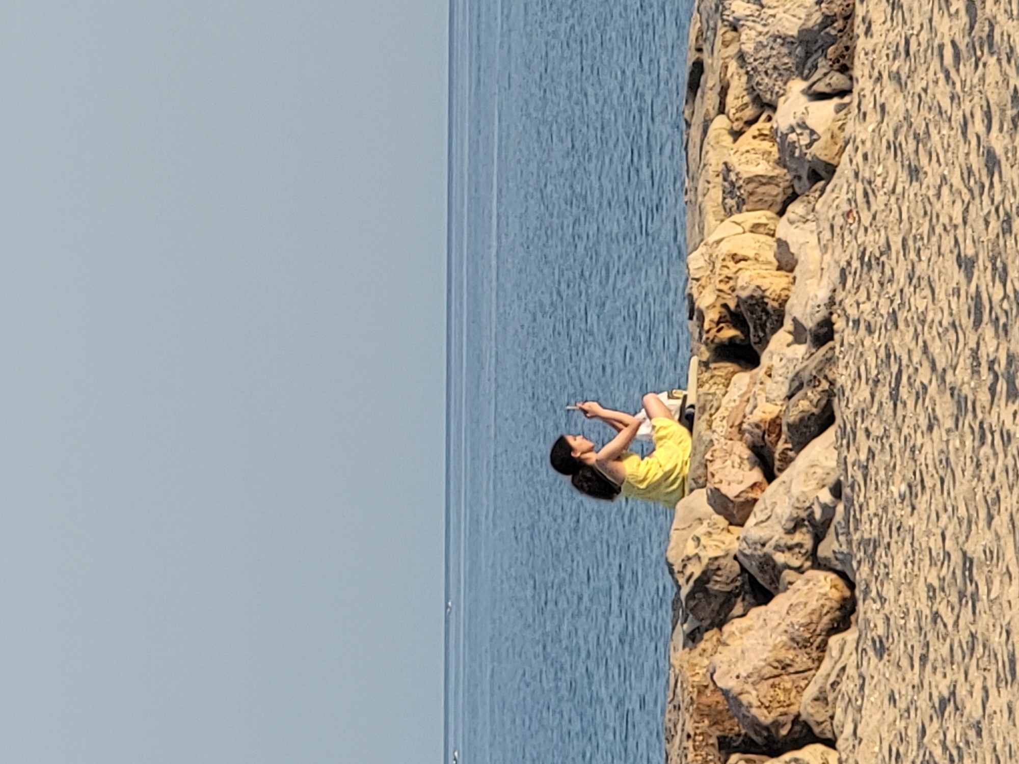 A young woman with dark, curly hair tied into a ponytail, wearing a yellow dress and white socks, sits on a rocky breakwater at the beach in Quarteira, Portugal. She is holding a smartphone in her hands and appears to be taking a picture or video, looking down at the screen. Her legs are crossed, and a white bag is placed beside her. The breakwater is made of large, rough-hewn rocks. Beyond the breakwater, the blue ocean stretches out to the horizon, with a clear, pale sky above. The foreground shows the sandy beach.