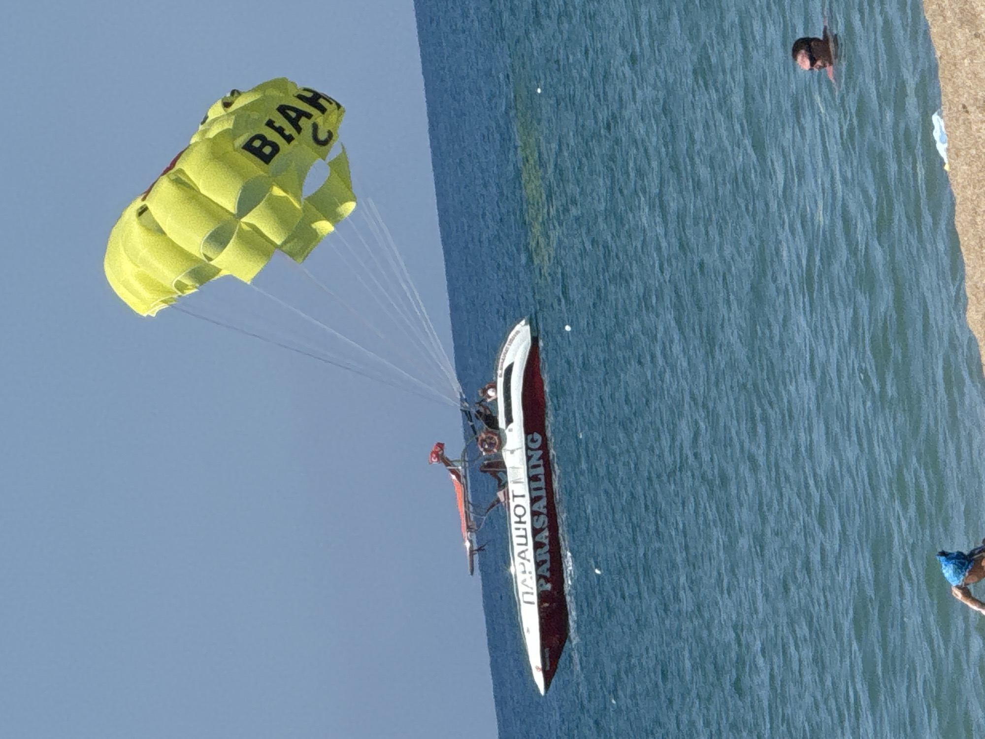 The image captures a parasailing activity on a sunny day at a beach in Manavgat, Turkey. A yellow parachute, bearing the word "BIAH," is aloft, tethered to a red and white parasailing boat that is moving through the turquoise water. The boat has "PARASUT" and "PARASAILING" written on its side. Two individuals appear to be on the boat, and another person, wearing sunglasses, is swimming in the sea in the foreground. In the lower part of the image, a person in a blue swim cap is partially visible, wading in the shallow water near the sandy beach. The scene is set against a clear blue sky and the rippling surface of the sea.