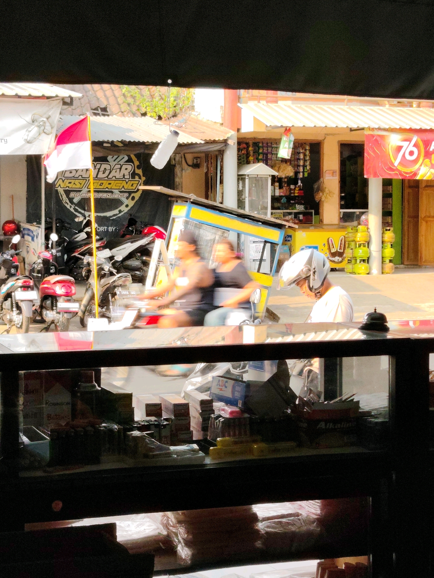 The image captures a street scene in Dampit, Indonesia, with a focus on a food stall and its surroundings. In the foreground, a glass display case reveals shelves stocked with various goods, including what appear to be cigarettes and other packaged items. Beyond the display case, a yellow and blue food cart is visible, with two people blurred due to motion, suggesting they are either riding or operating the cart. One person, wearing a white shirt and a helmet, is seated and looking down. Behind the food cart, a banner with Indonesian text, possibly advertising "Nasi Goreng" (fried rice), is prominently displayed along with an Indonesian flag. Several motorcycles are parked in front of the stall, indicating a common mode of transportation in the area. The background shows a row of buildings with awnings and signage, typical of a marketplace or commercial street. The overall impression is of a busy, everyday scene in a local Indonesian town.