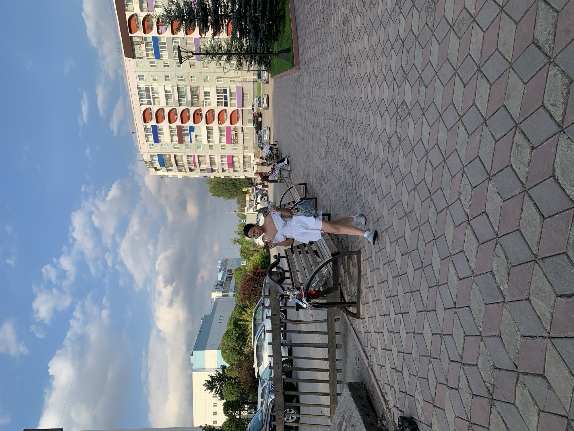 A young woman in a white dress walks along a patterned sidewalk in Prokopyevsk, Russia. She is holding a phone to her ear. Behind her, several people are sitting on a park bench, and a row of cars is parked along a street. In the background, there is a multi-story apartment building with colorful balconies and a large shopping center. The sky is partly cloudy, with the sun shining through. The area appears to be a public space, possibly a plaza or a pedestrian walkway, adjacent to a parking lot and residential buildings. The architecture of the apartment building suggests a Soviet-era or post-Soviet design. The sidewalk's geometric pattern is a prominent feature of the scene.
