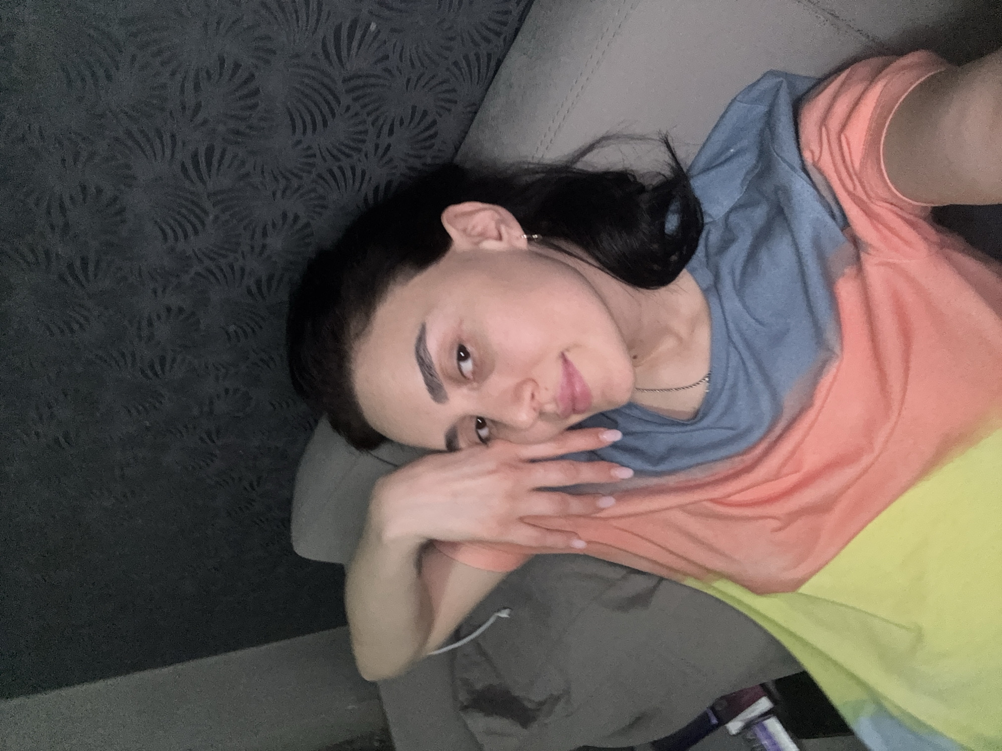A woman is lying on a gray couch, facing the camera. She has dark hair and is wearing a multi-colored shirt with blue, orange, and yellow sections. Her hands are near her face. The background includes a wall with a decorative pattern. The location cannot be determined solely from the image. There are no recognizable landmarks or identifying features to confirm the city of Tbilisi, Georgia.