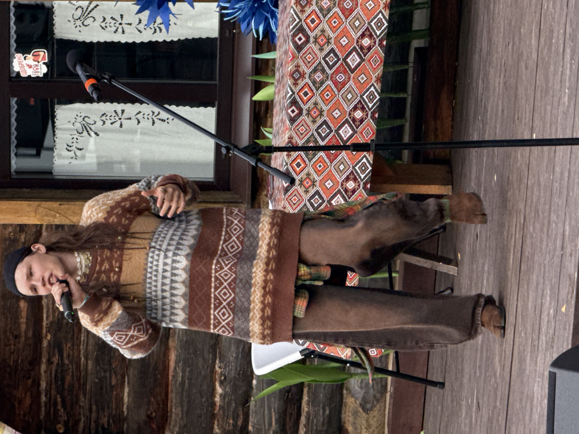 This media file features a young person with long brown hair, wearing a black beanie and a patterned sweater, holding a microphone and singing. The sweater has intricate geometric patterns in brown, white, and gray. The person is standing on a wooden floor, with a wooden wall and a window behind them. The window has white curtains with decorative patterns and is partially covered by a colorful, patterned fabric. Blue decorative elements are visible above the curtains. To the right of the person, there is a microphone stand and a black speaker. The scene appears to be outdoors, possibly at a festival or performance, given the microphone and the rustic wooden setting. The lighting suggests it is daytime. There are no strong cues for weather. The overall mood seems to be one of performance or entertainment. A small sticker with Cyrillic text, "добро твое" ("your good"), is visible on the window. The location context is Старые Быги, Russia.