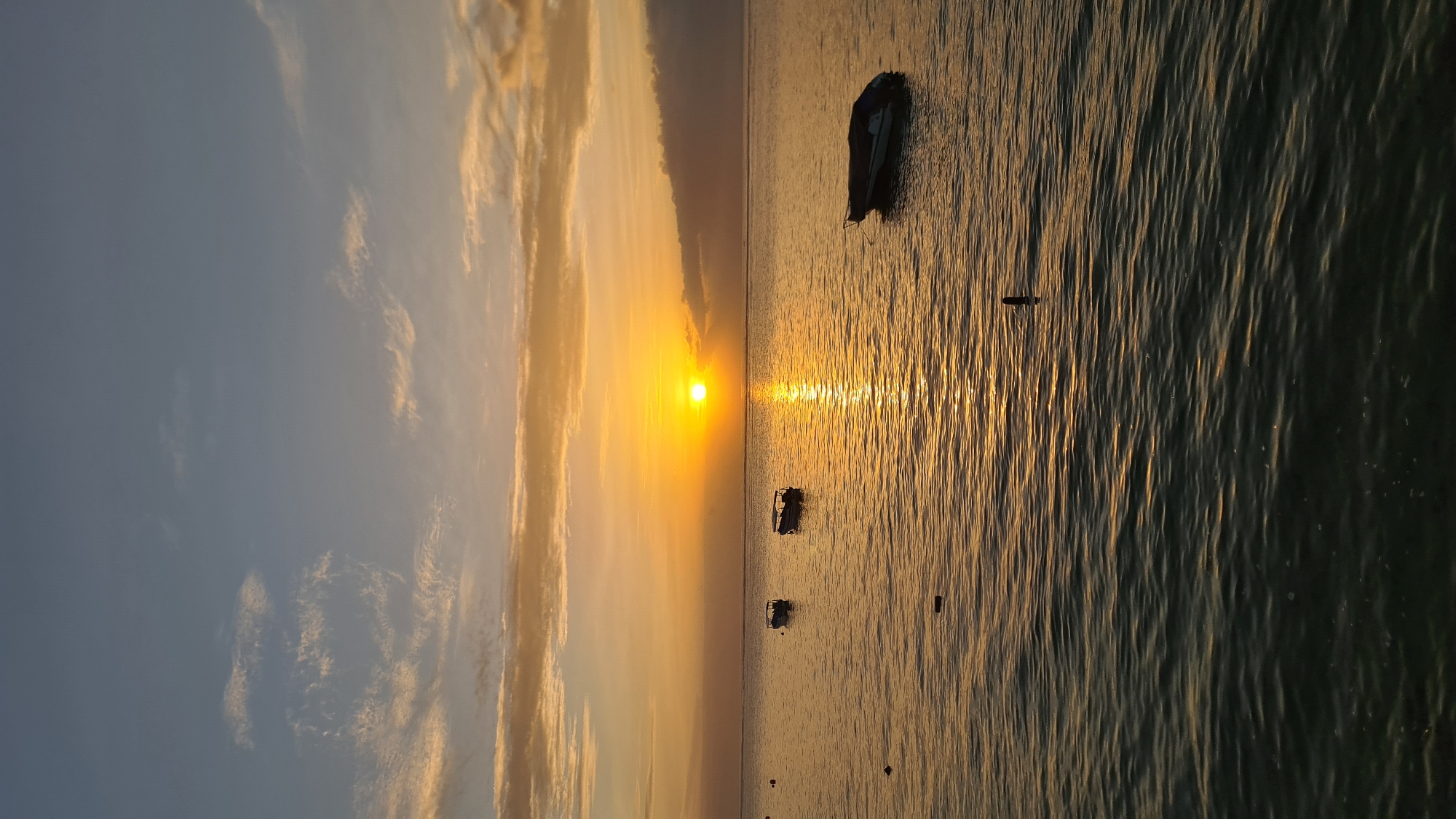 The image depicts a serene ocean sunset, likely in a coastal area of Denpasar, Indonesia. The sky is ablaze with warm hues of orange, yellow, and gold, transitioning into a soft blue at the top. Wispy clouds are scattered across the horizon, catching the last rays of the sun. The sun itself is a bright, radiant orb low on the horizon, casting a shimmering golden path across the water.
Several small boats are visible on the calm sea. Two boats are positioned closer to the left side of the frame, appearing to be moored or anchored. Further to the right, another boat is seen, its silhouette distinct against the glowing water. The water surface is rippled, reflecting the colors of the sky and creating a textured, dynamic effect. In the foreground, the water is darker, suggesting a deeper perspective or a shift in lighting. A few dark, small objects, possibly buoys or markers, are scattered in the water. The overall atmosphere is peaceful and tranquil, capturing a moment of natural beauty at the end of the day.