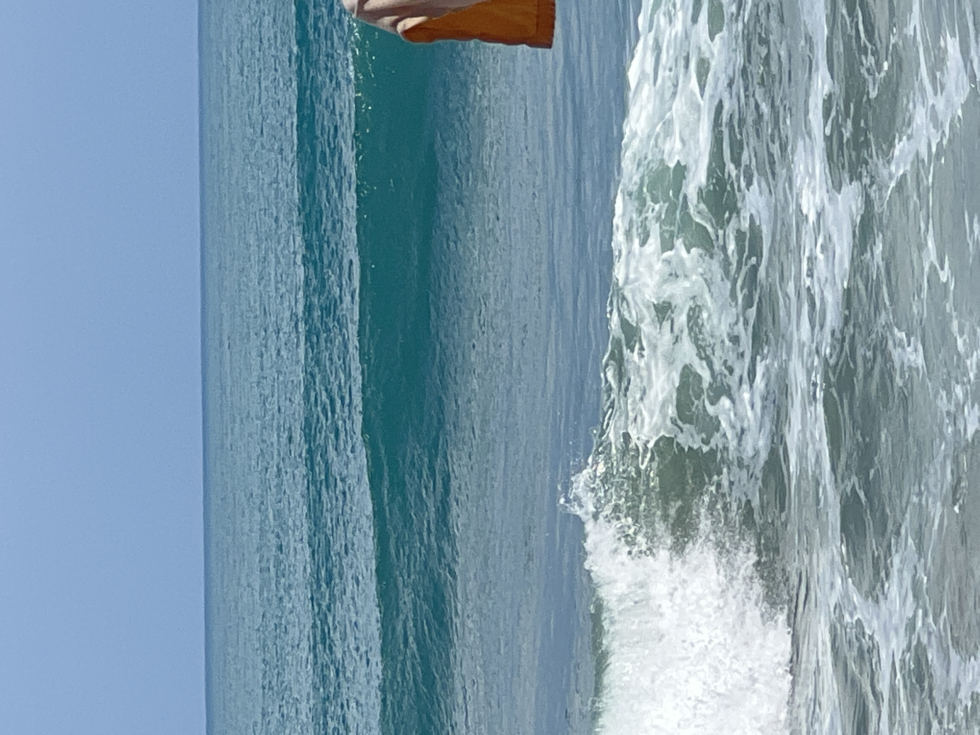 A wide shot of the ocean off the coast of Kuta, Indonesia, shows gentle waves rolling in under a clear blue sky. The water is a clear turquoise, with white foam visible where the waves are breaking closer to the shore. In the right side of the frame, a person wearing an orange garment is visible from the waist down, standing in the water. The person appears to be holding something in their hands. The scene is peaceful and serene, with the sounds of the ocean likely to be the only sounds present. The water is calm and inviting, and the sky is a clear, cloudless blue. The overall impression is one of tranquility and natural beauty.
