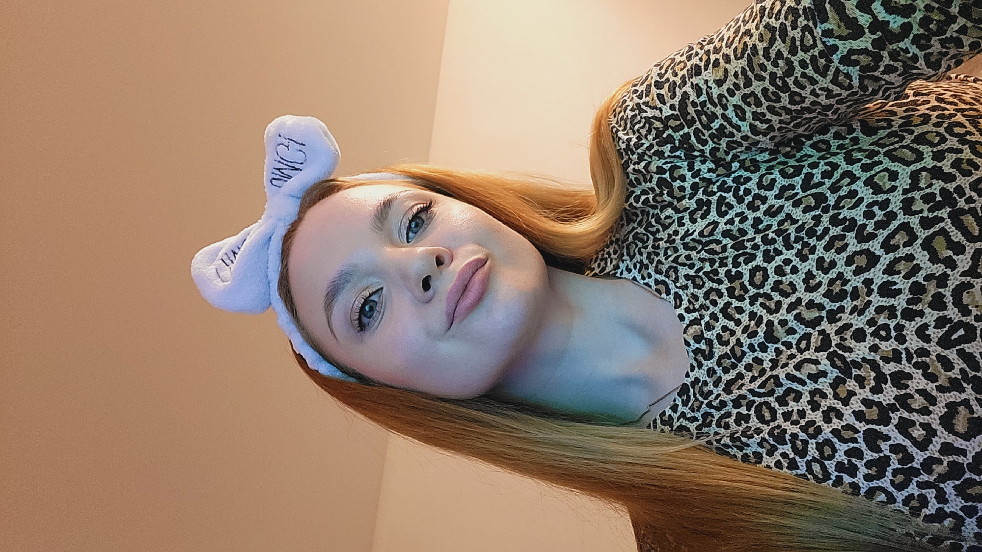 A young woman with light skin and light brown hair poses for a selfie. She is wearing a leopard-print top and a white headband with bunny ears that has the word "OMG!" embroidered on it. The background is a plain, light tan wall. The photo appears to have been taken indoors, potentially at a residence. There is no information in the image to confirm the location in Chaikovsky, Russia.