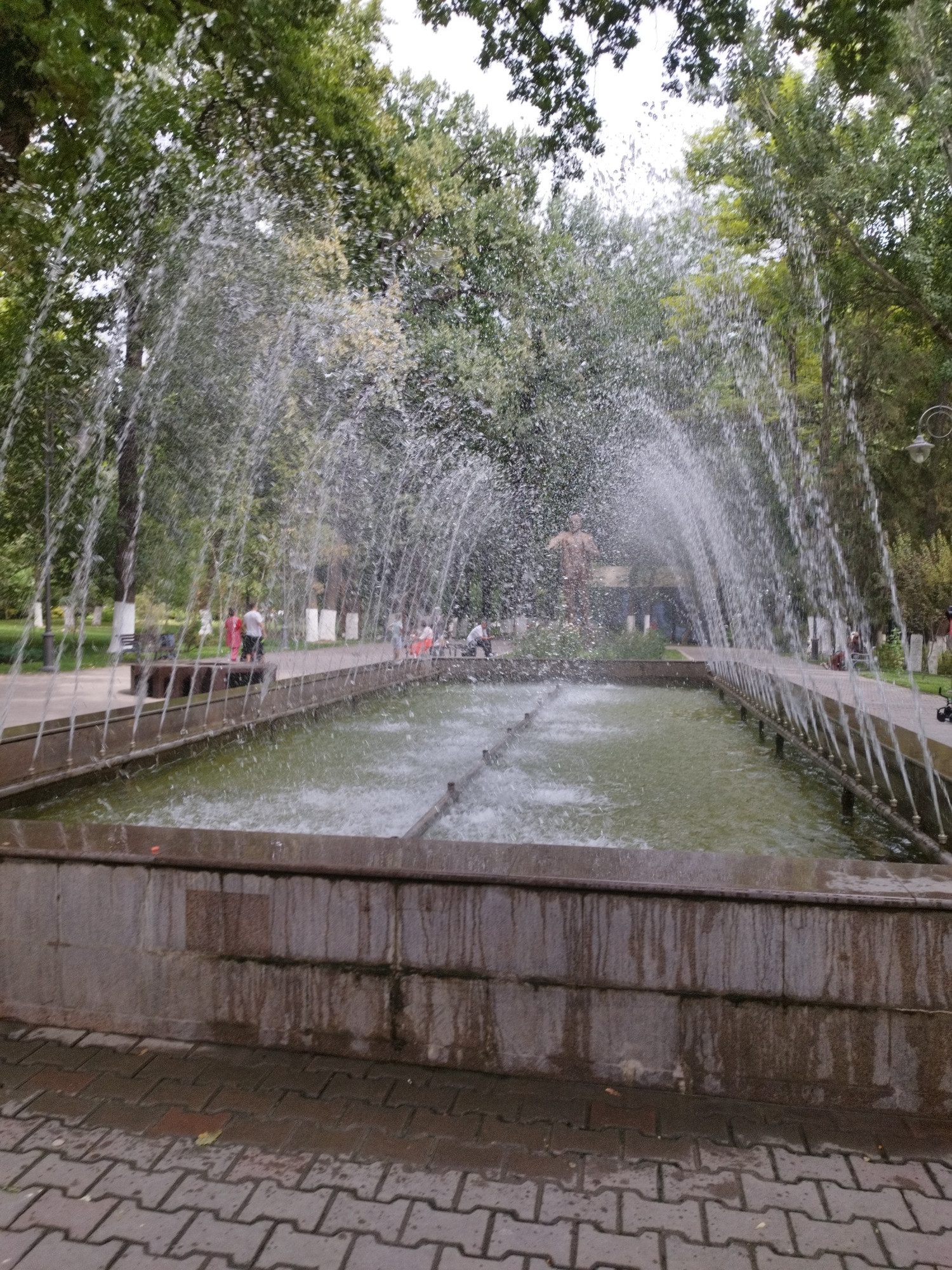 The image depicts a fountain in a park-like setting, likely within the city of Shymkent, Kazakhstan. The fountain is the central focus, with water spraying in arching streams over a rectangular pool. The pool is surrounded by a stone basin, which shows signs of wear and water staining. Beyond the fountain, a paved pathway runs parallel to it, lined with trees on one side. Several people are visible in the background, some walking along the path and others sitting on benches. A statue of a person is visible behind the fountain on the left. The overall environment suggests a public park, possibly in an urban area, with lush green trees and paved walkways. The water from the fountain is creating a misty effect, obscuring some of the finer details in the background.