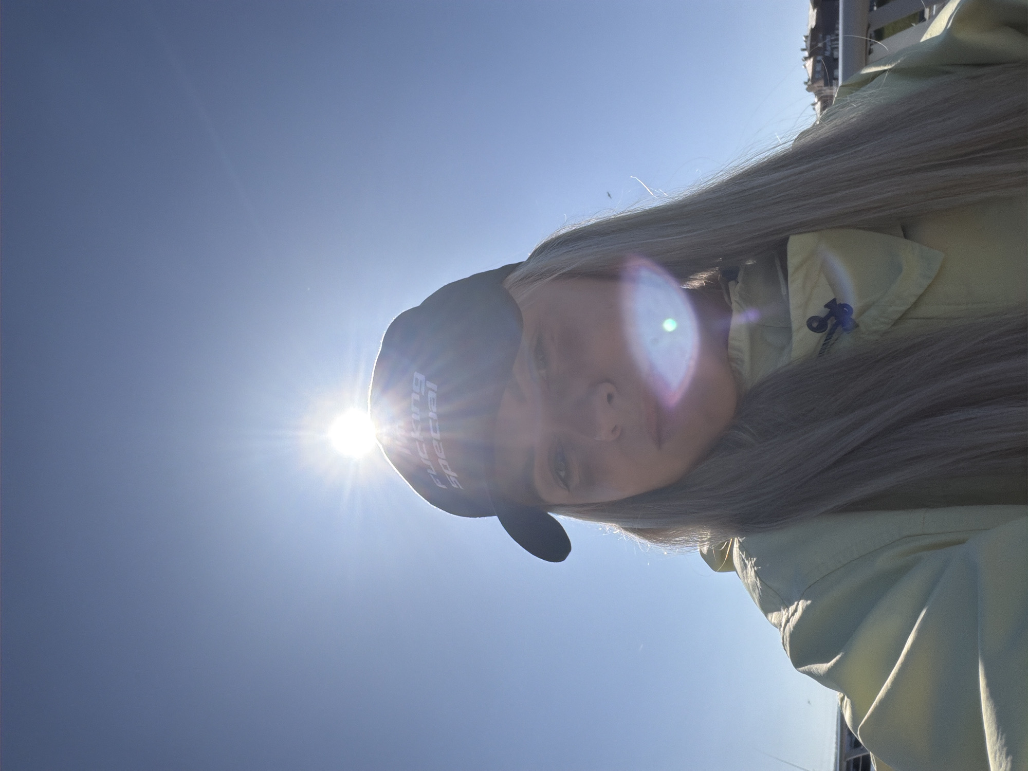 A person wearing a black cap with "FUCKING SPECIAL" printed on it and a light green jacket is positioned at an angle against a bright blue sky. The sun is directly behind the person's head, creating a lens flare effect. Long, gray hair is draped over the person's shoulder and jacket. The location is undetermined, and there is no information given to specify the location is in the city of Votkinsk, Russia. No action, interaction, or additional subjects are observed.