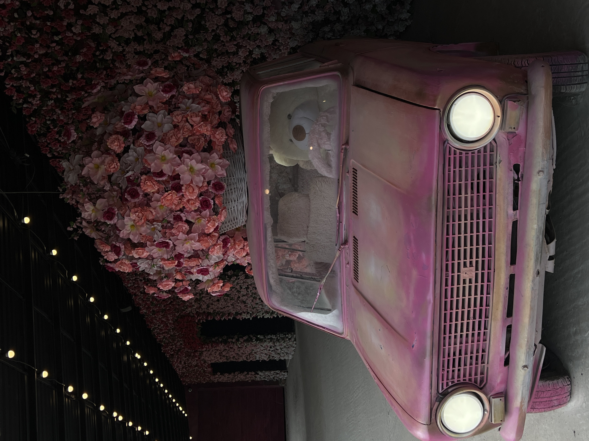 The media file presents a whimsical and colorful display, likely within an indoor or covered setting. A vintage-style car, painted in a distressed pink with areas of fading and wear, is a prominent subject, oriented vertically in the frame as if on its side. Its round headlights are illuminated, casting a bright glow. The car features a vertical slatted grille, and a portion of a pink-painted tire is visible at the top right.
Inside the car, a large, fluffy white teddy bear with a light brown snout is visible through the windshield and passenger-side window, appearing to look outwards.
Above and surrounding the car is a dense arrangement of artificial flowers, primarily in shades of pink, peach, and white, creating a lush, floral canopy or wall. A large wicker basket brimming with similar flowers is positioned next to the car's passenger side.
To the left, a dark wall or surface is adorned with multiple strands of warm, glowing string lights, arranged diagonally across the frame. The overall lighting is dim, enhancing the atmospheric and dreamy quality of the scene. The setting appears to be an artistic installation or themed environment, possibly a cafe or retail space. There is no visible text. The image's location is Ternopil, Ukraine.