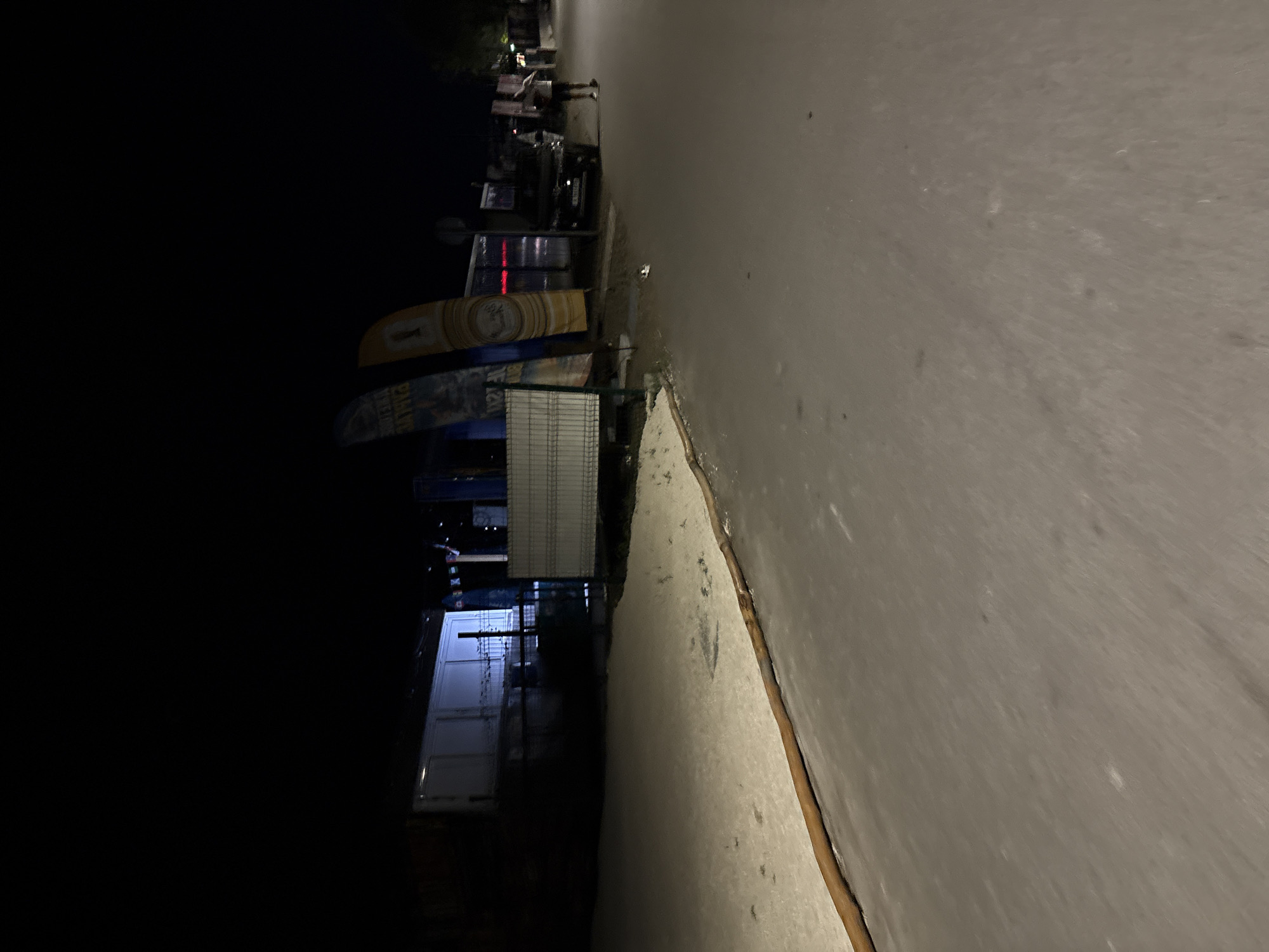 The image depicts a nighttime scene on a street in Burgas, Bulgaria. In the background, a long, white, modular building or container unit is visible, with some fencing in front of it. Adjacent to this structure are several tall, vertical banners, two of which are clearly visible and appear to be promotional flags. One flag is blue with white lettering, and another is yellow with circular graphics. A dark-colored vehicle is parked on the right side of the street. A person, seen from the side and slightly from the back, is standing near the vehicle, appearing to be wearing shorts and a t-shirt. They are facing away from the camera. The street itself is paved and appears wet, reflecting some of the ambient light. A sandy or gravelly area runs along the side of the street, bordered by a dark, elongated object, possibly a log or a pipe, on the edge of the paved surface. The overall lighting is dim, characteristic of nighttime conditions, with some artificial light sources illuminating the building and banners.