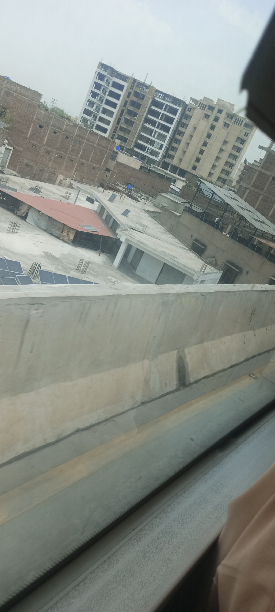 The image depicts a cityscape, likely in Alam Khan Kalay, Pakistan, with a focus on construction and urban development. In the background, a multi-story building is under construction, showing exposed concrete framework and window openings. Adjacent to it is another completed building with a similar architectural style. The foreground reveals rooftops of various structures. One rooftop features a red-tiled canopy covering an area with construction materials and scaffolding. Another prominent feature is a bank of solar panels installed on a rooftop. Several other smaller buildings with flat, concrete roofs are visible, some with what appear to be small utility structures or vents. The overall scene is characterized by a mix of ongoing construction and established urban elements, under an overcast sky. The perspective suggests the photo was taken from an elevated position, possibly from a moving vehicle or a higher building, as indicated by the angled concrete barrier in the immediate foreground.