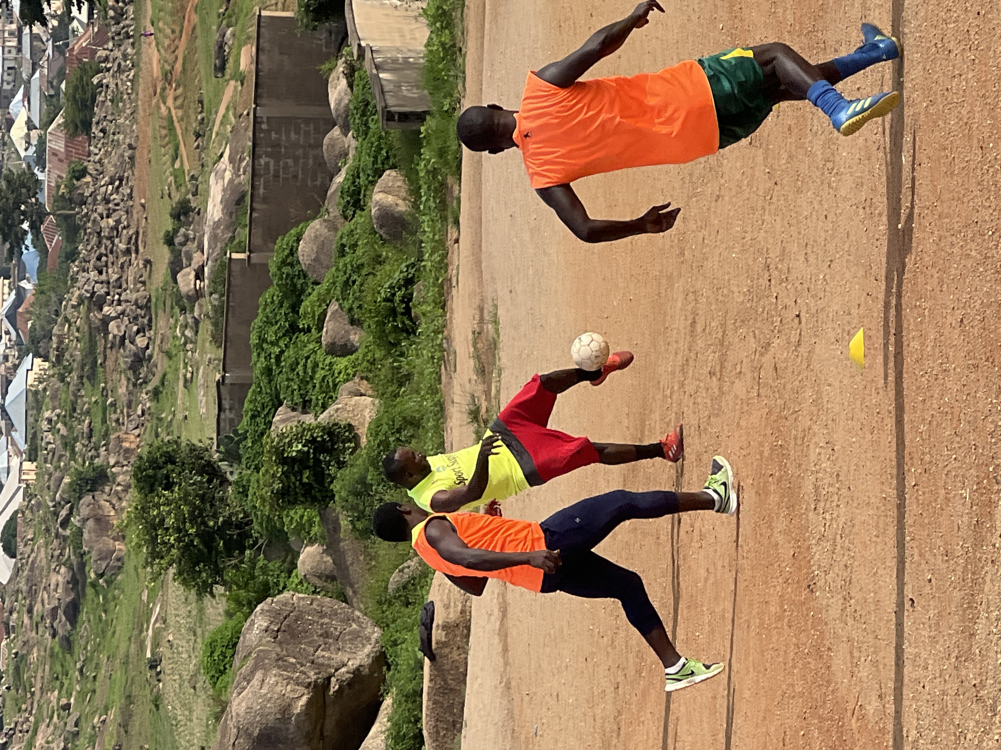 In Jos, Nigeria, three individuals are engaged in a soccer game on a dirt field. One player, wearing a yellow sleeveless shirt and red shorts, is kicking a white soccer ball. Another player, in an orange sleeveless shirt and dark blue pants, is running, with his left foot raised. The third player, also in an orange sleeveless shirt and green and yellow shorts, is running away from the viewer, facing right. The background reveals a hilly, rocky landscape with scattered buildings and vegetation, characteristic of the environment in Jos. The lighting suggests it is daytime, likely afternoon, with shadows cast on the ground.