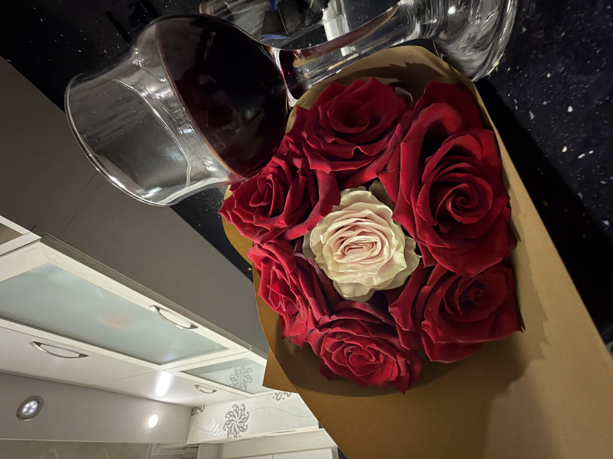 A high-angle, close-up shot, likely taken in Pazardzhik, Bulgaria, captures a bouquet of roses and a wine glass filled with red wine. The scene is set indoors, appearing to be a kitchen or dining area, illuminated by artificial light suggesting evening or night.
The primary subjects are a bouquet of seven roses and a large wine glass. The bouquet features six vibrant deep red roses encircling a single pale pink or cream rose at its center. The roses are wrapped in plain, natural brown paper. Beside the bouquet, a clear stemmed wine glass is tilted on its side, holding a generous amount of dark red wine, with reflections visible on its surface.
Both objects rest on a dark, speckled countertop, possibly granite or a similar material. In the background, modern white upper kitchen cabinets with frosted glass panels and sleek metal handles are visible. Recessed lighting fixtures cast light onto the scene, and a subtle decorative white floral pattern can be seen on a surface or wall below the cabinets.
There is no specific activity depicted, but the combination of roses and wine strongly implies a romantic gesture, a celebration, or a moment of relaxation. No visible text is present in the image.