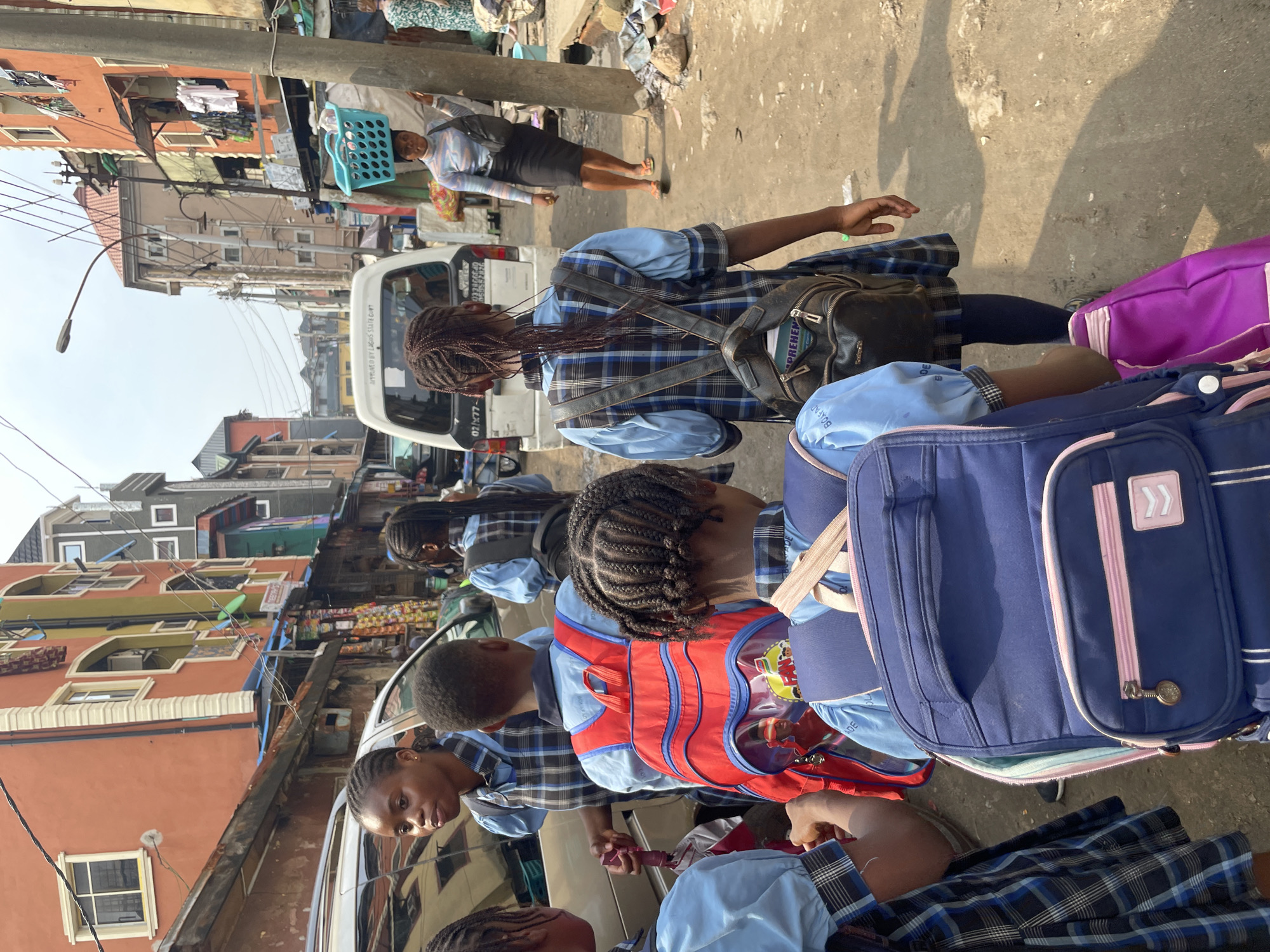 In a crowded Lagos, Nigeria street scene, several schoolgirls in blue and black plaid uniforms and backpacks stand near a white minivan. The van has a registration plate indicating it's "Approved by Lagos State Govt." and bears the partial number "02/X77-". The backpacks are a mix of blue and purple. The girls appear to be interacting, with one looking towards the others. Above and behind them, the street is lined with buildings in varying states of repair and color, including yellow, orange, and green. Overhead power lines and other urban infrastructure are visible. In the background, a woman carries a blue plastic laundry basket. Litter is strewn on the ground.