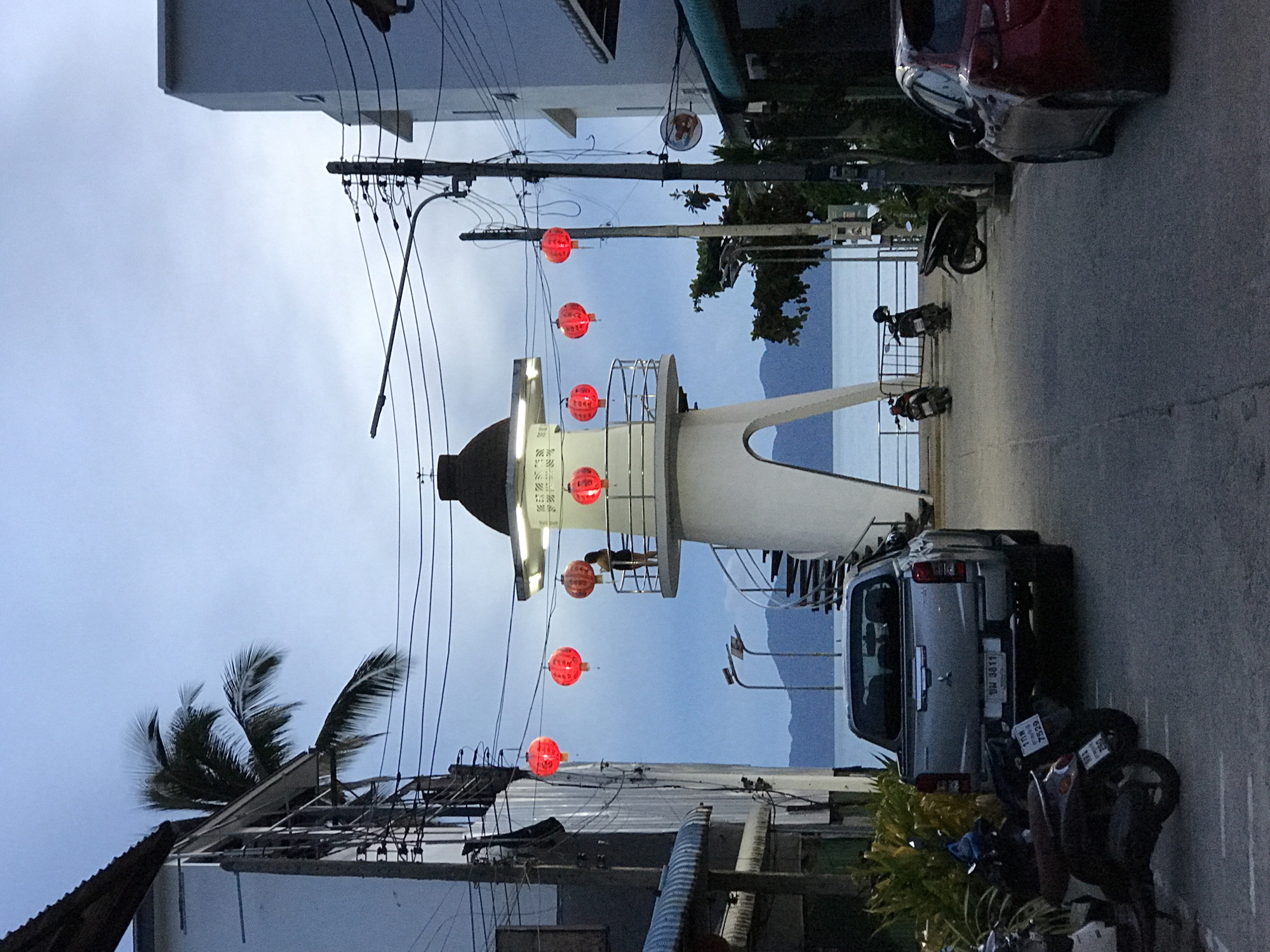 The image depicts a street scene in Baan Mae Nam, Thailand, at dusk. A prominent white lighthouse-like structure stands in the middle of the street, adorned with red lanterns. Overhead power lines crisscross the scene. Buildings line both sides of the street. A silver pickup truck is parked on the right side of the street, along with several motorcycles. In the background, the sea and distant mountains can be seen. A palm tree is visible on the left side of the image. A red car is parked on the other side of the street and another motorcycle is parked right on the side of the street.