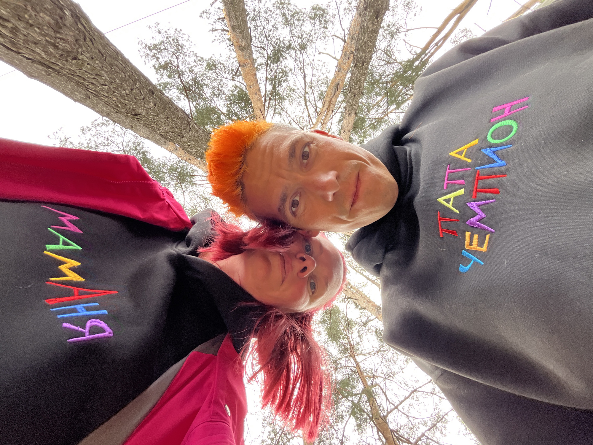 An upward-angled outdoor photograph captured in Chaikovsky, Russia, features two individuals with brightly colored hair positioned head-to-head at the bottom of the frame. The person on the left, appearing female, has vibrant red hair and wears a black hoodie with a visible pink lining or trim. Embroidered on her hoodie, facing the viewer, are the Cyrillic letters "МАМА" (Mama), rendered in green, yellow, red, blue, and purple thread. Her gaze is directed slightly upwards towards the camera, with a neutral expression. The person on the right, appearing male, has vivid orange hair and wears a dark grey or black hoodie. Embroidered on his hoodie, oriented similarly, are the Cyrillic letters "ПАПА УЕМТМОН" (Papa Yemtmion), also in a multi-colored thread sequence including red, yellow, light blue, purple, green, and orange. His eyes are directed straight at the camera, with a subtle, calm expression. Both individuals' heads are tilted towards each other, creating an intimate pose. The background consists of numerous tall, slender tree trunks and bare branches reaching towards a bright, uniformly overcast sky, indicating an outdoor, possibly forested, setting during a season of sparse foliage.