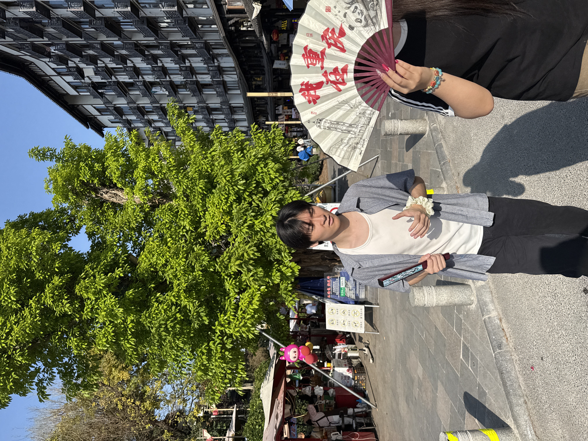 A young man stands on a paved street in Shapingba District, China, holding a closed fan. A woman behind him holds an open fan with Chinese characters and a panda illustration. A large tree with green foliage is to the left. A building with many windows is in the background. A vendor stand with various items is partially visible.