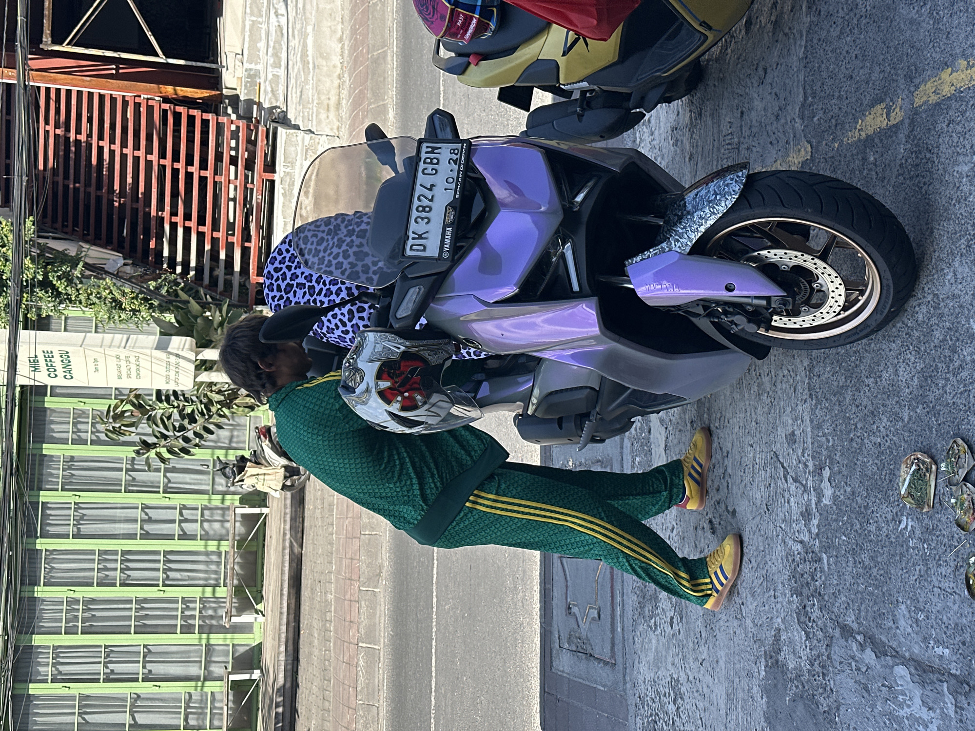 A man in a green and yellow tracksuit is standing next to a purple Yamaha scooter. The scooter's license plate reads "DK 3824 CBN." The man is holding a motorcycle helmet. The scooter is parked on a sidewalk next to a street. In the background, there is a building with green window frames. The building has a sign that reads "MIEL COFFEE CANGGU." The location is North Kuta, Indonesia, based on the signage.