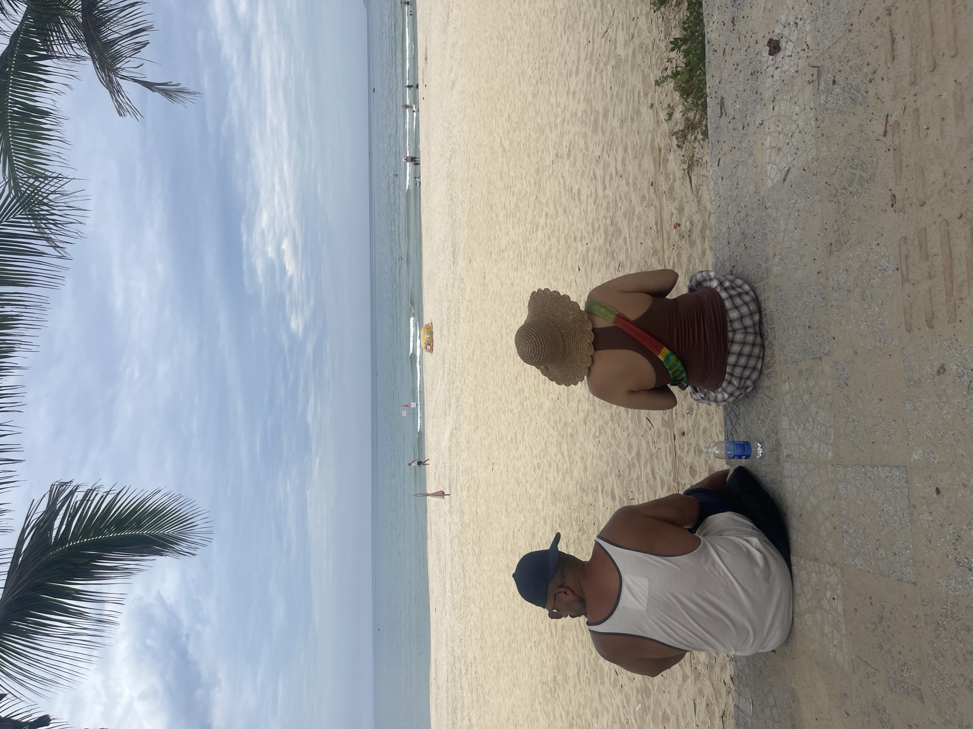 A couple is seated on a concrete ledge overlooking a sandy beach and the ocean in Đà Nẵng, Vietnam. The man on the left wears a white tank top and a dark baseball cap. The woman on the right wears a brown tank top and a straw hat, with a colorful strap across her shoulder. A water bottle sits between them. The beach is sparsely populated with a few people in the distance, some wading in the water. Palm fronds frame the top of the image. The sky is overcast with clouds. The scene appears to be a relaxed day at the beach.