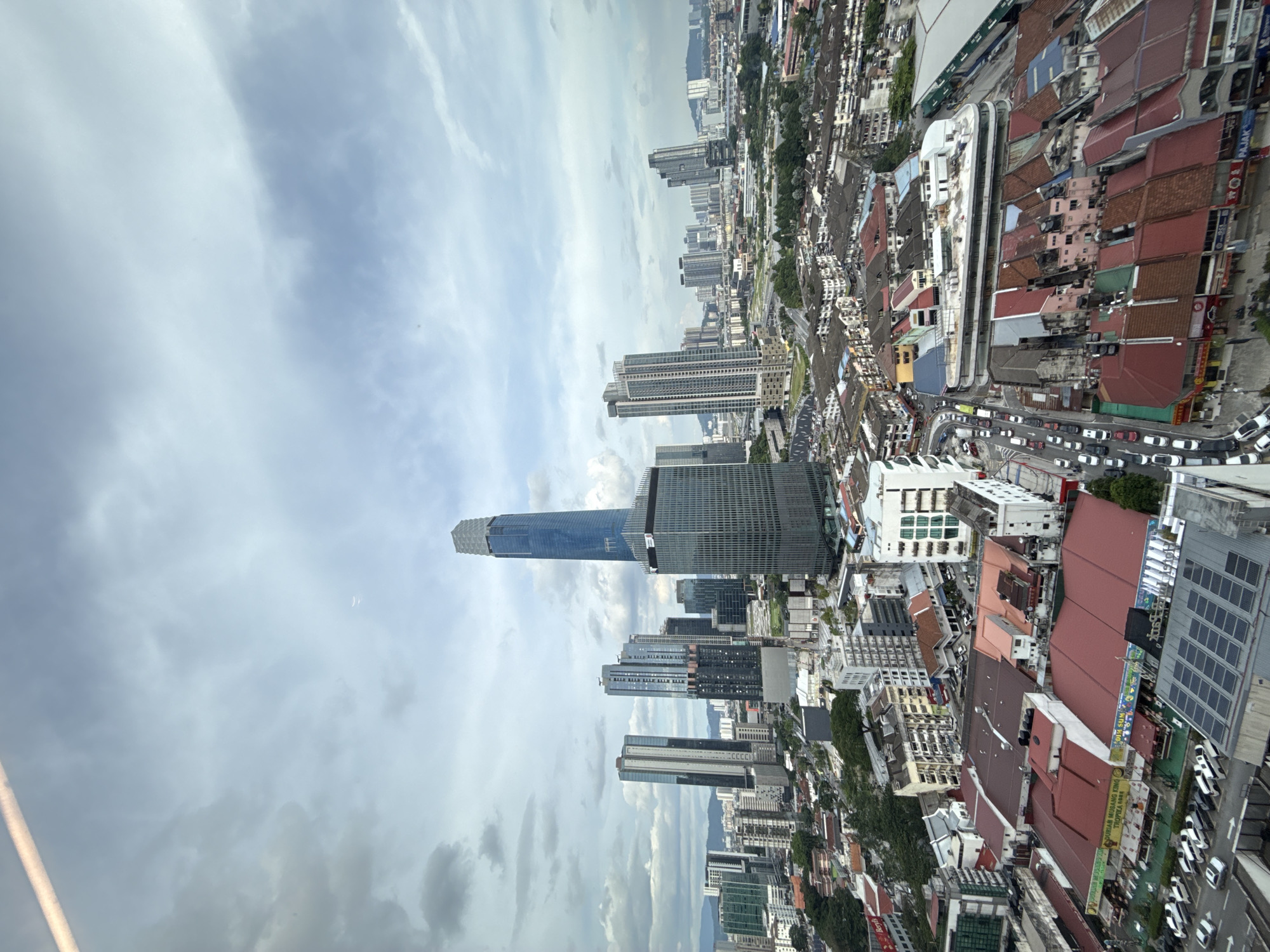 The image is a high-angle, slightly rotated shot of Kuala Lumpur, Malaysia. In the foreground, a densely packed urban area with a mix of low-rise buildings with red-tiled roofs and taller white or light-colored buildings is observed. Cars line the streets, suggesting traffic. In the midground, there's a cluster of modern skyscrapers, including the prominent Merdeka 118, with its distinctive tapered design. The background contains a cityscape extending into the distance, with more buildings partially obscured by atmospheric haze or clouds. The sky is cloudy, with varying shades of gray and white. The overall scene portrays a bustling city with a blend of older and modern architecture under overcast conditions.