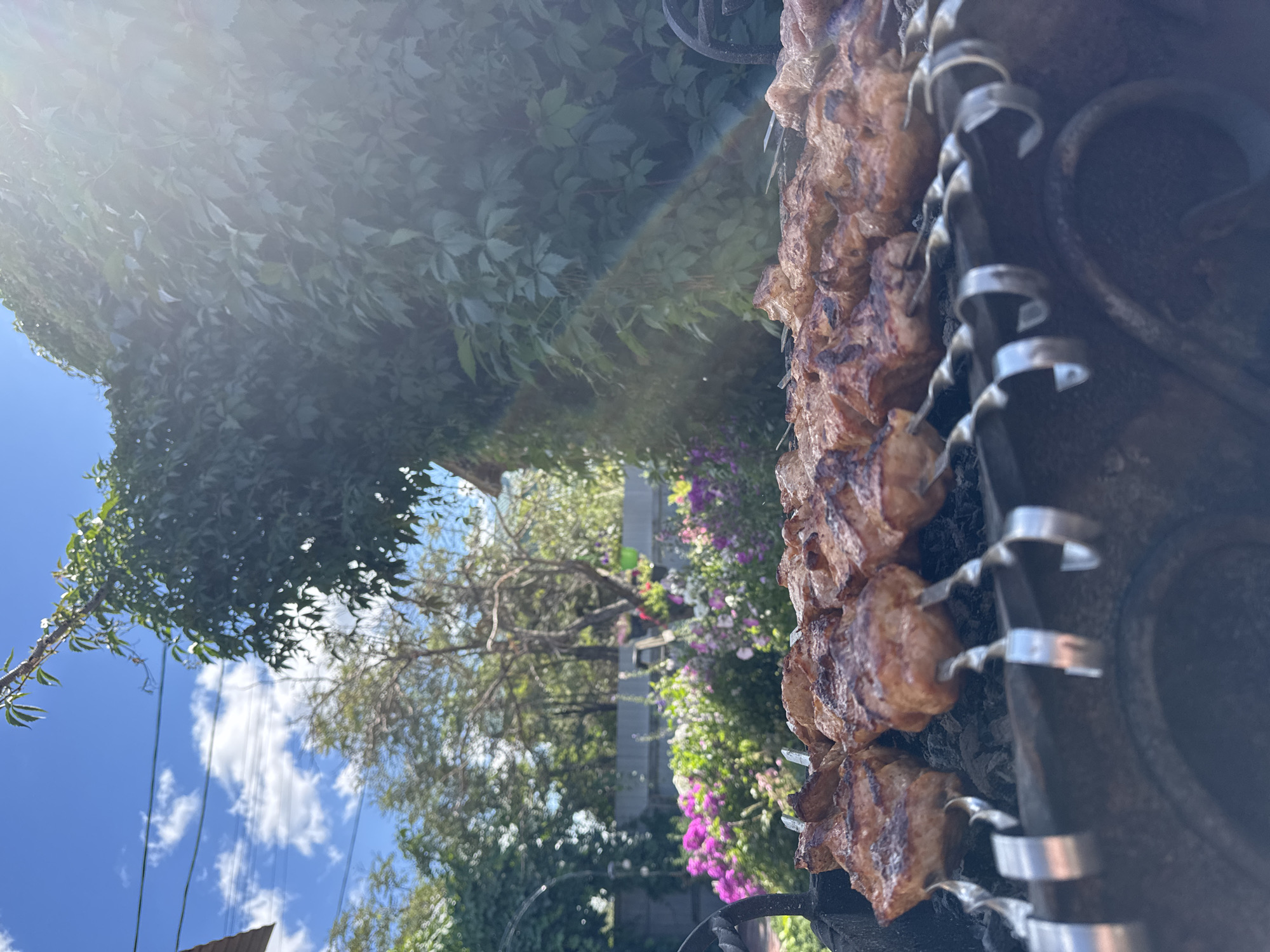 The image shows a close-up view of meat skewers cooking on a charcoal grill in an outdoor setting. The meat is browned and appears to be in the process of being grilled. The grill is made of metal and is filled with hot coals. In the background, there is a lush green garden with flowers. The sky is blue with white clouds, and sunlight is streaming down, creating lens flare in the upper right portion of the image. The scene suggests a summer day spent outdoors. The location is specified as Prokopyevsk, Russia. There are no people visible in the image.