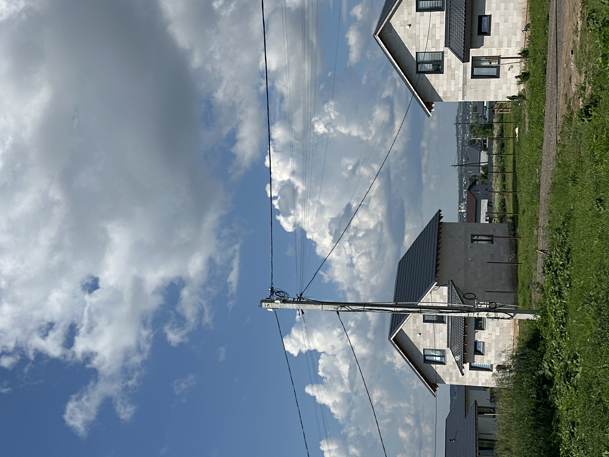 The image captures a scene in a semi-rural area, possibly a developing residential zone, with two unfinished houses constructed from light-colored blocks and dark roofs. The houses are situated on a slight incline, with a dirt path in the foreground leading towards them. Lush green grass and some scattered vegetation are visible in the foreground and around the houses. A concrete utility pole with overhead wires stands prominently in the center-left of the frame, interrupting the view of the sky. The sky itself is a dynamic mix of bright blue and large, billowy white and grey clouds, suggesting a day with changeable weather. In the background, a distant, hazy urban or suburban landscape with more buildings and greenery is visible. There are no people or animals present in the image. The overall impression is one of a construction site or a newly developing neighborhood under a dramatic sky.