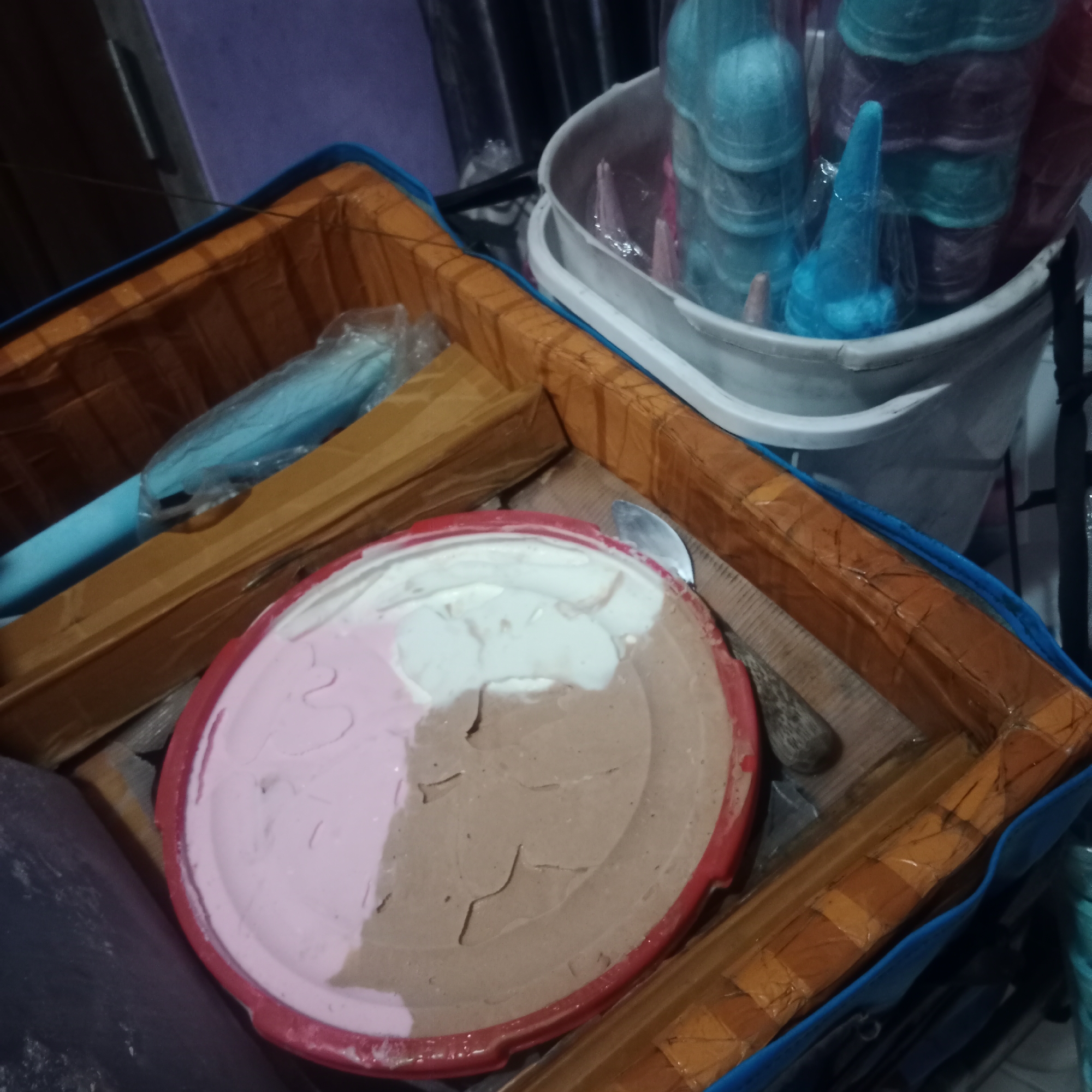 In the city of Tegowanu Kulon, Indonesia, a close-up shot captures an insulated cooler bag filled with frozen treats. The cooler bag, primarily blue with orange interior lining, holds a red round container of ice cream. This ice cream is tri-colored, with distinct sections of pink, white, and chocolate brown. A metal spoon rests partially within the ice cream container. To the left of the ice cream, a blue cylindrical object, possibly an ice pop mold, is visible, partially wrapped in plastic. In the background, a white bucket contains an assortment of blue and purple ice pops, many still in their plastic packaging, stacked vertically. The overall scene suggests a vendor's setup, likely for selling ice cream.