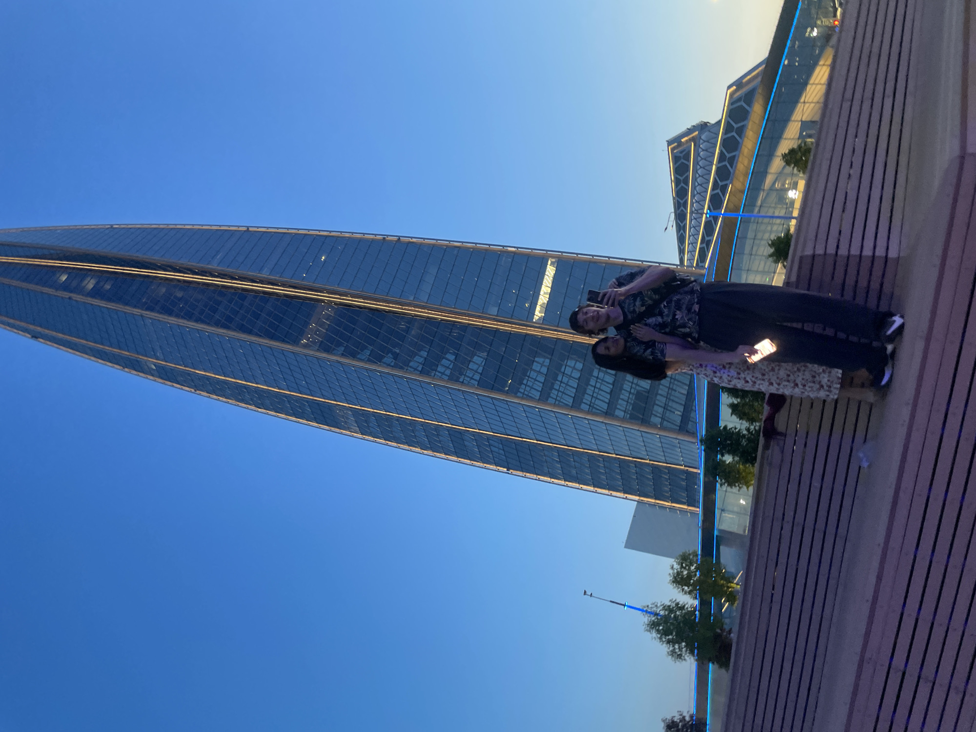 A couple is standing on a wooden structure, with a man taking a selfie of himself and the woman he is with. The man is wearing a dark t-shirt with a tropical pattern and dark pants. The woman is wearing a dress with a floral pattern. They are both holding phones. In the background, there is a modern, glass-covered skyscraper illuminated with yellow lights, which is the Lakhta Center in Saint Petersburg, Russia. To the right of the skyscraper is another contemporary building with a geometric design, also illuminated with blue and yellow lights. The sky is clear and blue, indicating it is either early morning or late evening. The wooden structure appears to be an outdoor seating area or a tiered platform.