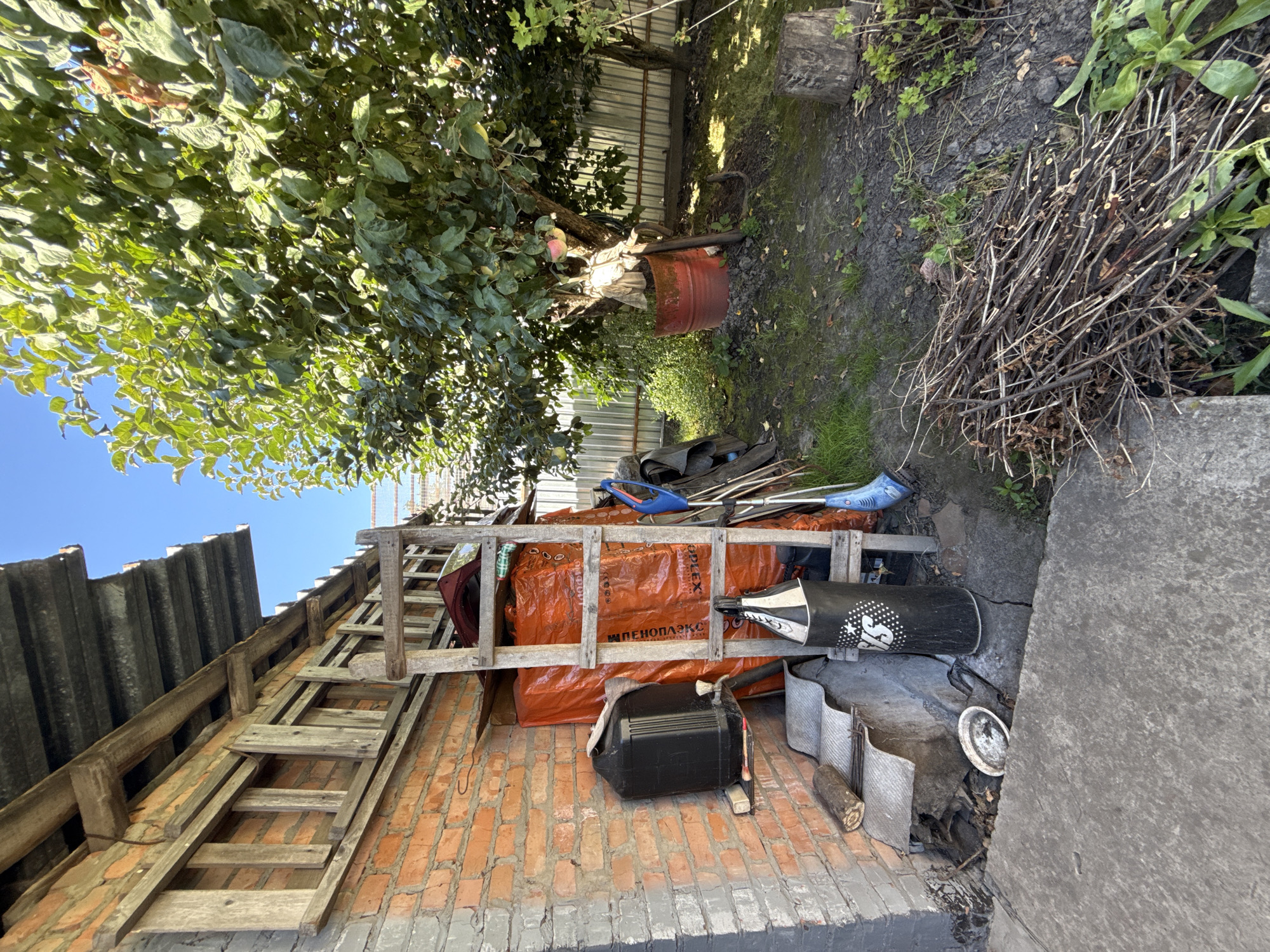 The image depicts an outdoor storage area next to a brick building, possibly in the city of Zheleznogorsk, Russia, given the context. A slanted metal roof covers part of the building and extends over a wooden ladder leaning against the wall. The brick wall is weathered. Against the wall, there is a large black plastic container with a spigot, resting on a metal stand. Adjacent to it, a black punching bag with white lettering is leaning. Behind these items, several large orange plastic-wrapped objects are stacked, supported by a wooden ladder structure. In the foreground, a concrete slab forms a pathway or ground surface. To the right, a tree with green foliage and some visible fruit stands in a red barrel. Several garden tools, including a broom and a hoe, are leaning near the tree. A pile of cut branches lies on the ground near the concrete path, and a tree stump is visible further to the right. The overall scene suggests a utilitarian outdoor space with various stored items and gardening implements. The lighting indicates it is daytime with clear skies.